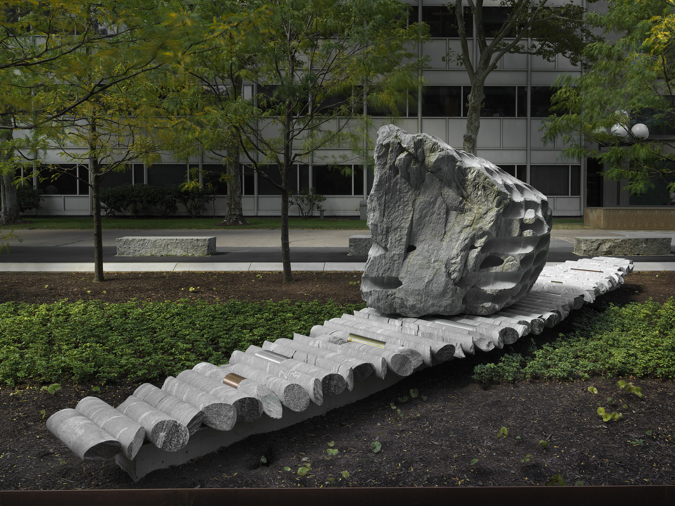 A large rock sculpture rests on a long strip of stone cylinders in a  greenspace. The rock has a series of holes removed from its core, with the  cylinders that were removed making up the strip it rests on. Trees, a pathway,  and a gray building reside in the background.