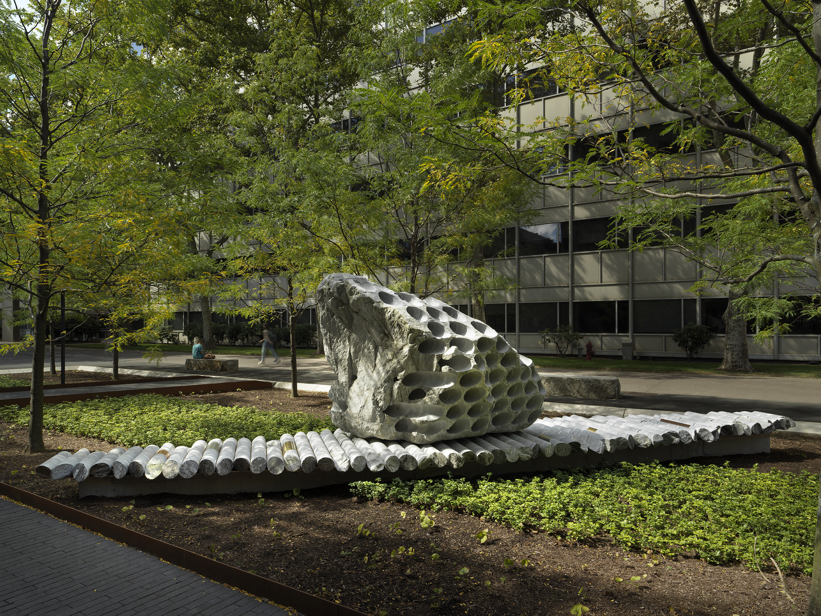 A large rock sculpture rests on a long strip of stone cylinders in a  greenspace. The rock has a series of holes removed from its core, with the  cylinders that were removed making up the strip it rests on. Trees, a pathway,  and a gray building reside in the background.