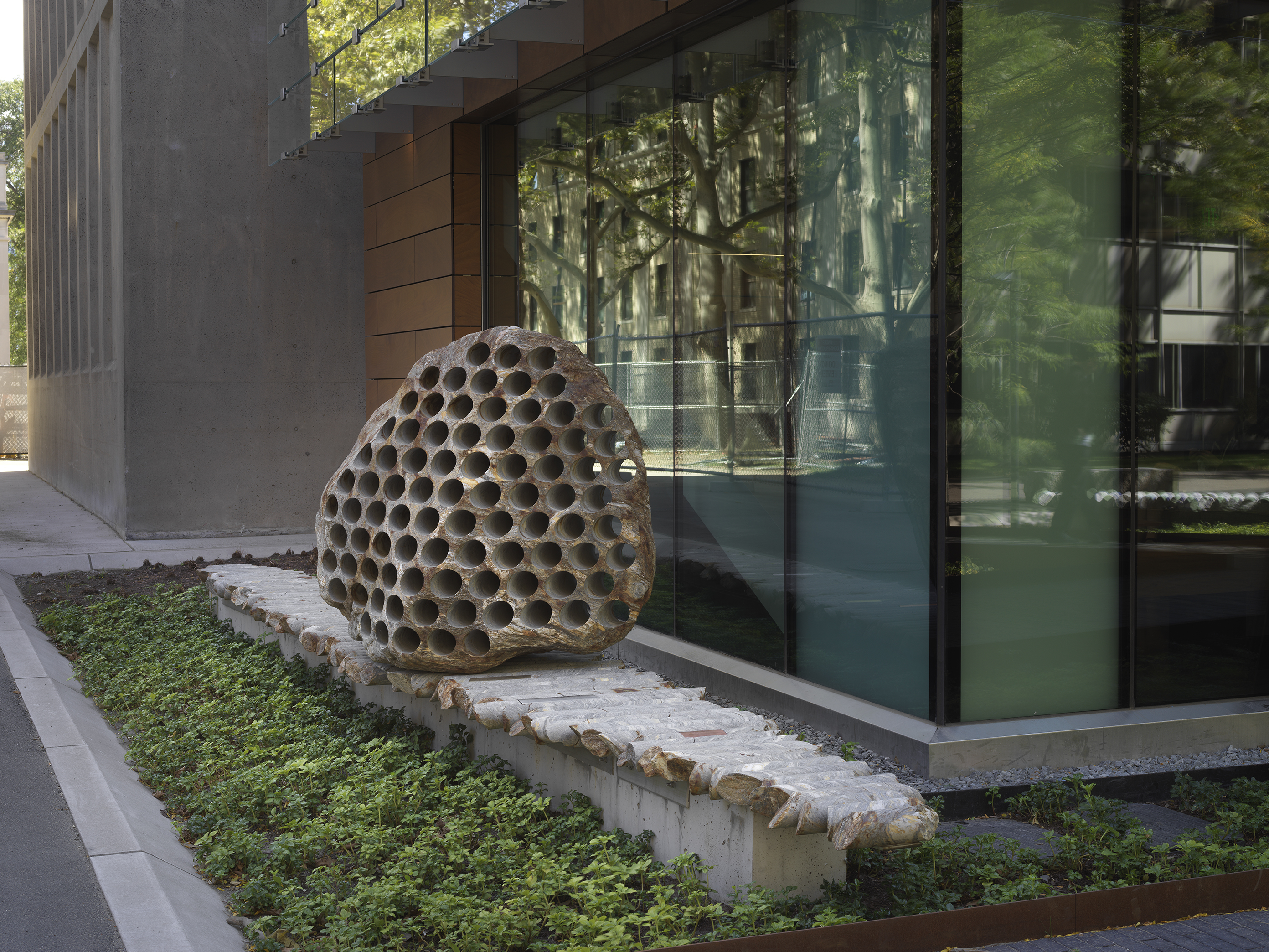 A large rock sculpture rests on a long strip of stone cylinders in a  greenspace next to the corner of a building. The rock has a series of holes  removed from its core, with the cylinders that were removed making up the strip it  rests on.