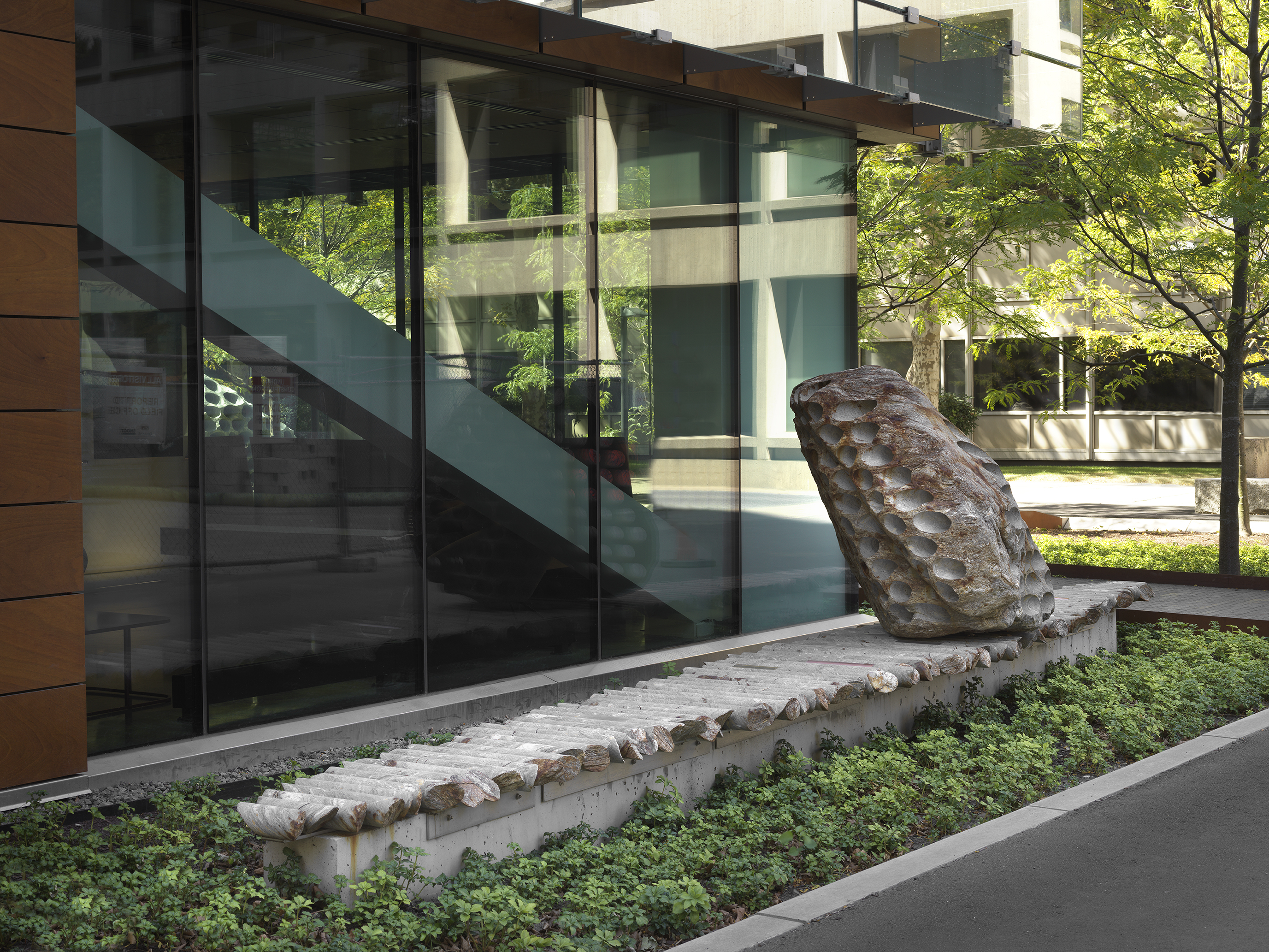 A large rock sculpture rests on a long strip of stone cylinders in a  greenspace next to the corner of a building. The rock has a series of holes  removed from its core, with the cylinders that were removed making up the strip it rests on.