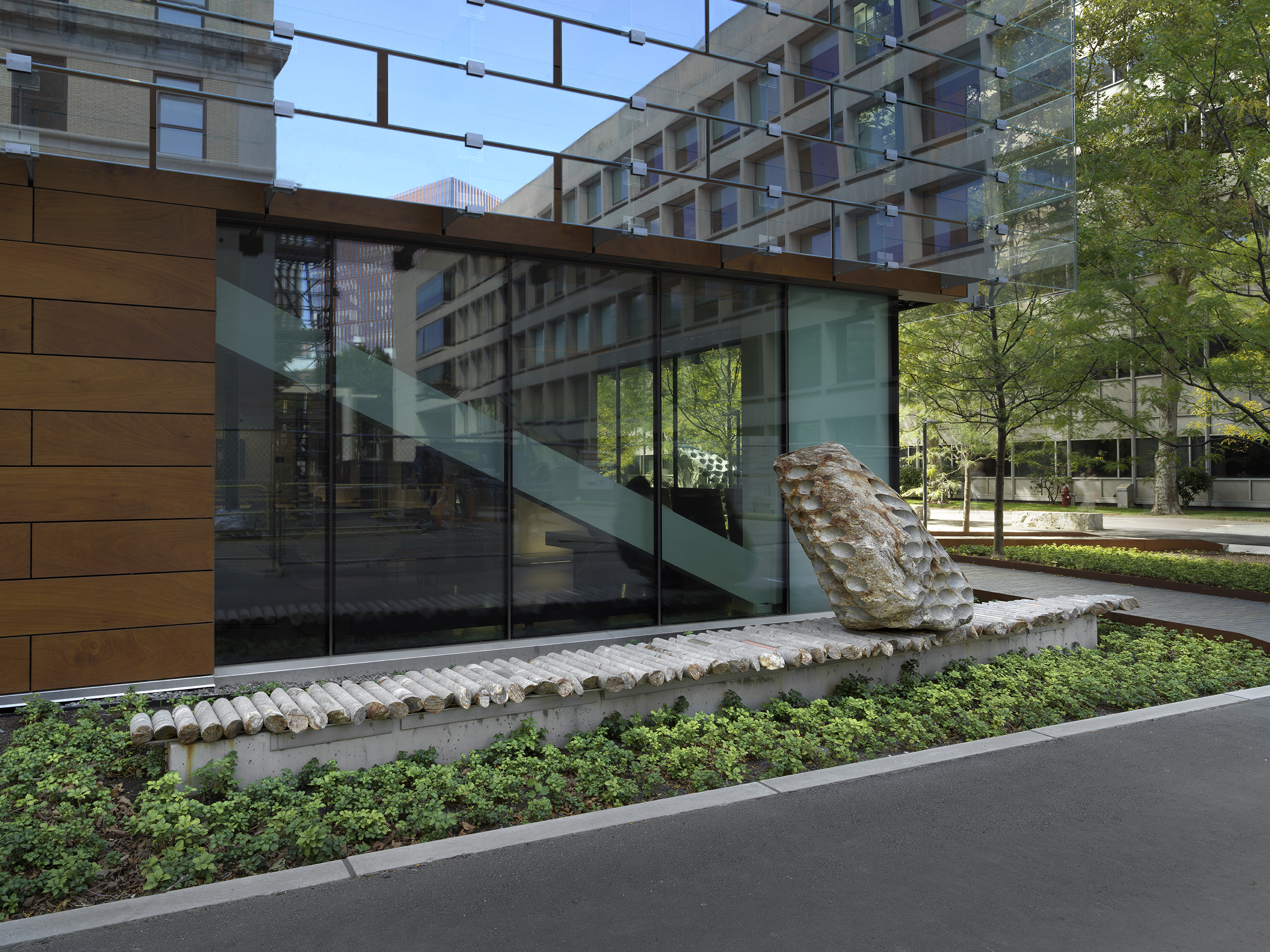 A large rock sculpture rests on a long strip of stone cylinders in a  greenspace next to the corner of a building. The rock has a series of holes  removed from its core, with the cylinders that were removed making up the strip it rests on. 