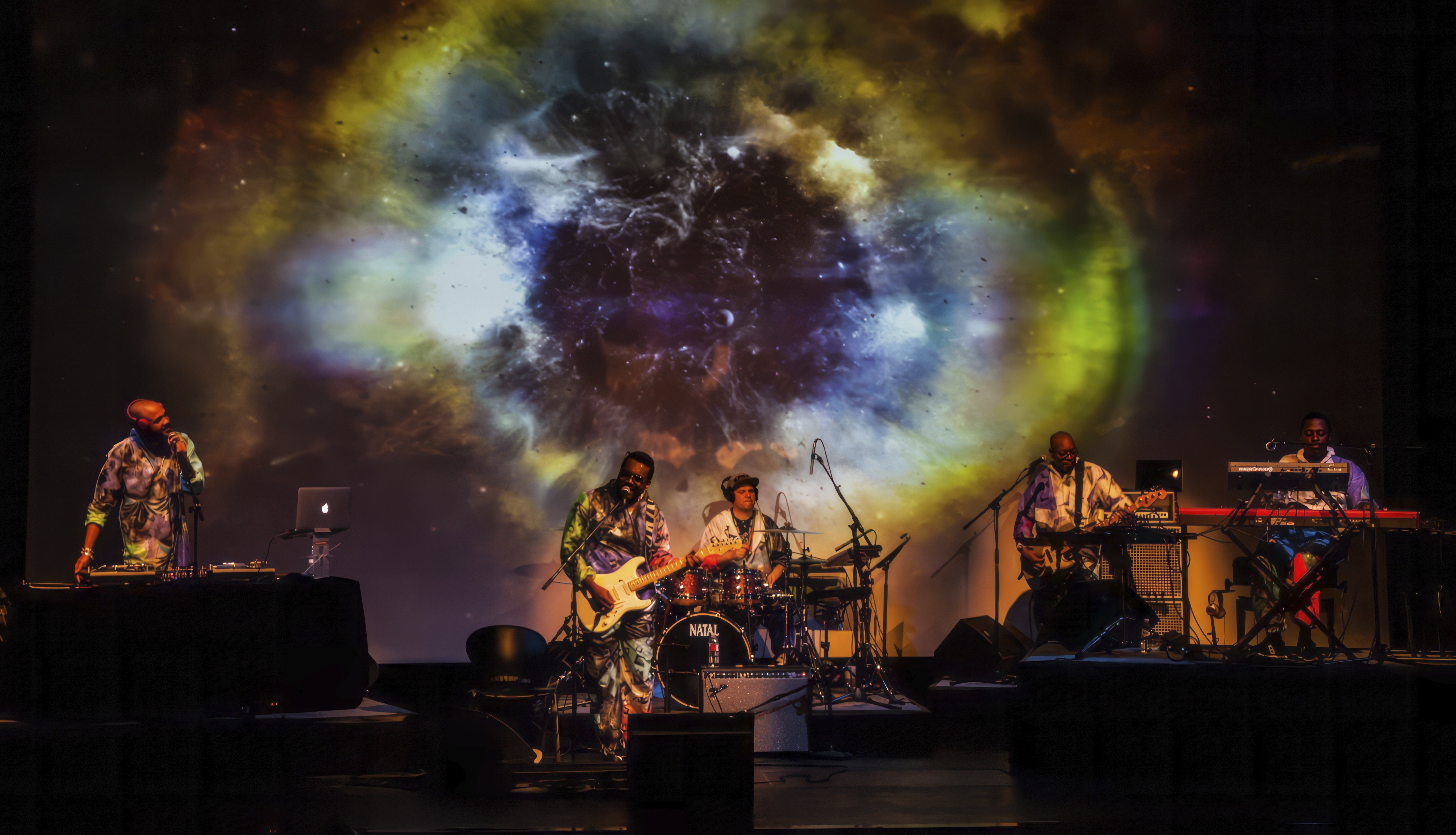 Image of a live musical performance, with five band members on stage in front of a projection of a galaxy.