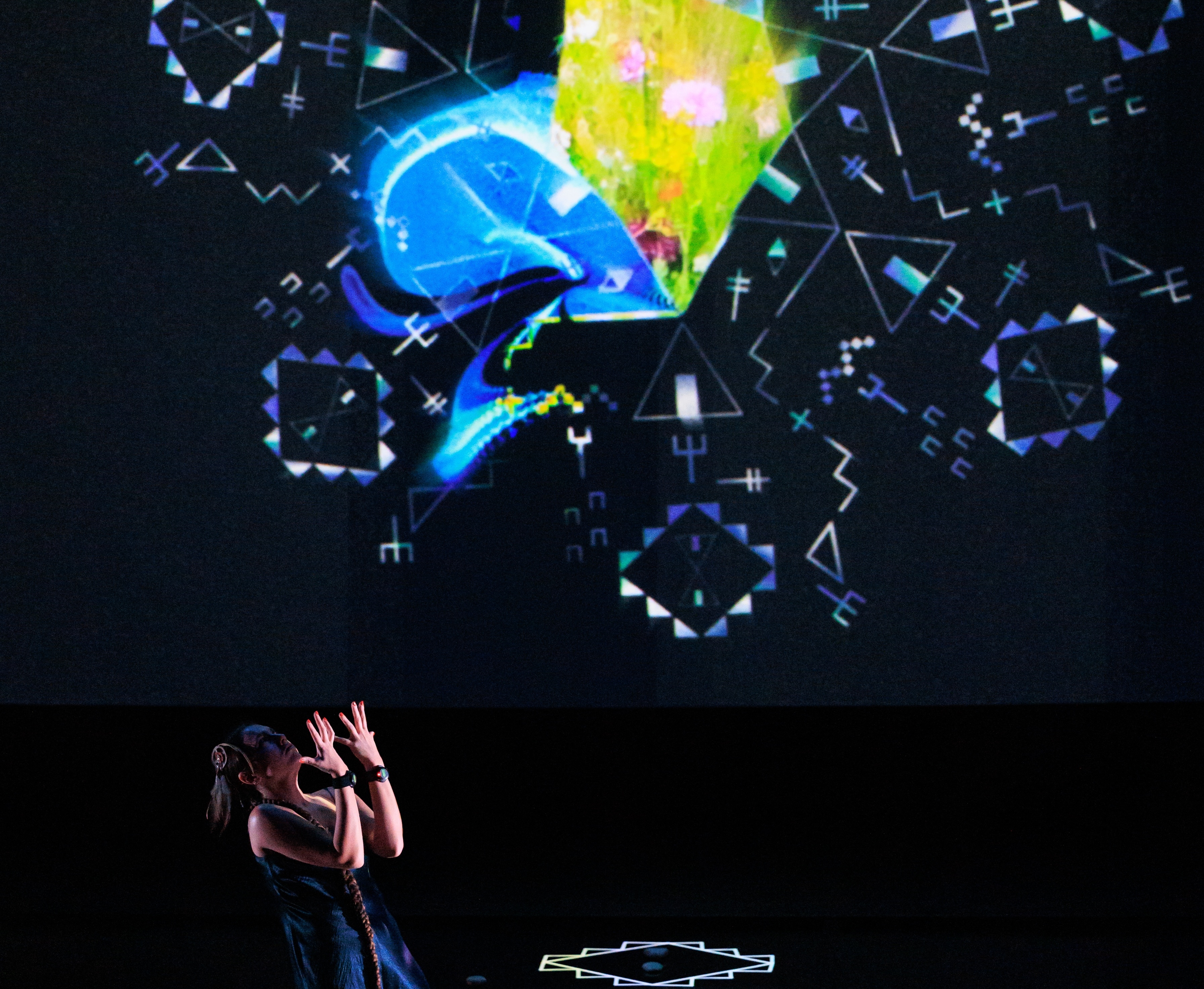Video still of a performance showcasing a woman spotlighted on stage with her head tilted up, arms raised and palms facing up. A brightly colored geometric pattern in the background is juxtaposed by a black wall.