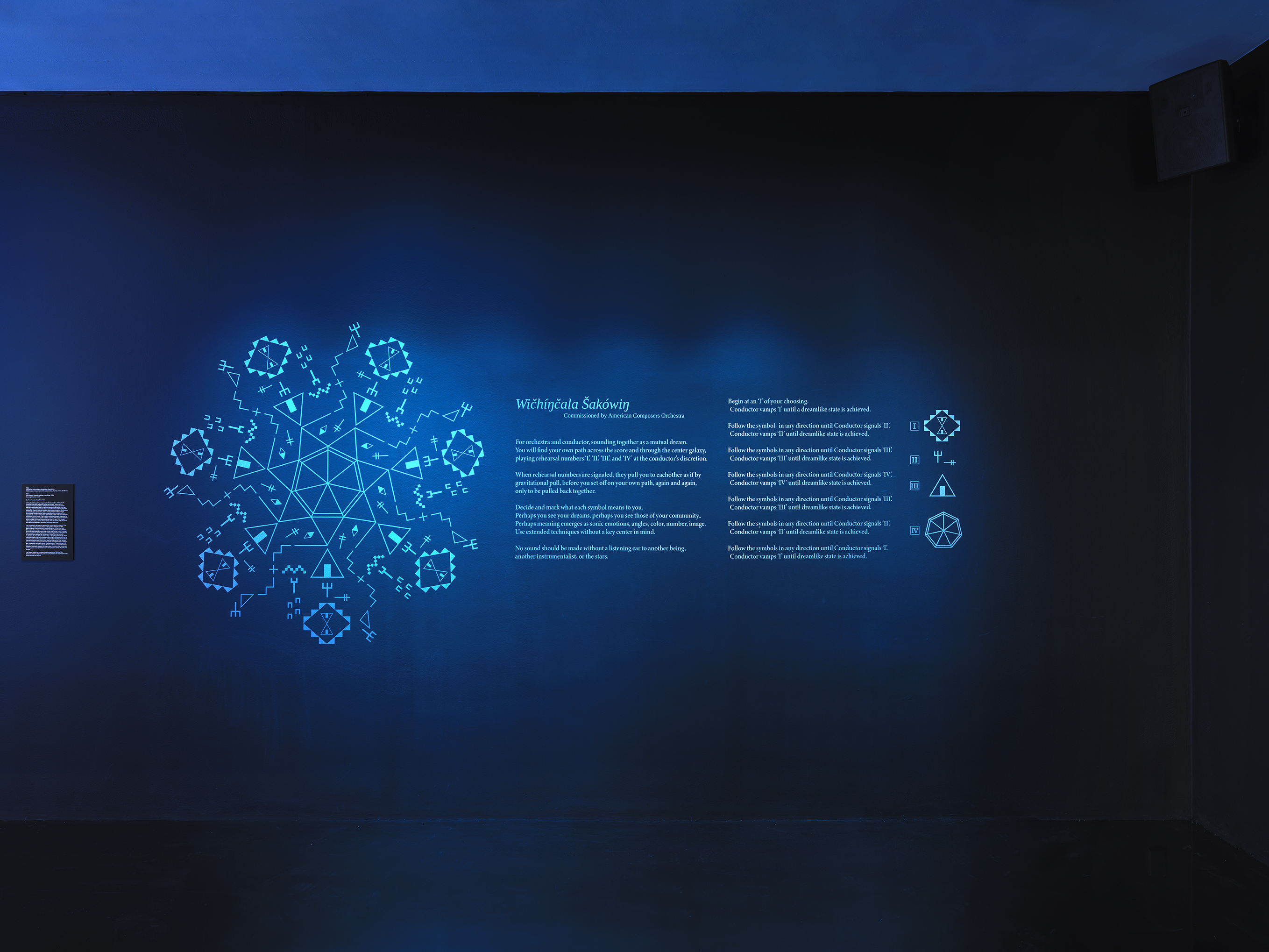 On a dark blue gallery wall, a glowing circle of geometric forms made up of‬ ‭ Lakhota characters is displayed. To its right, instructions for a score commissioned‬ ‭ by the American Composers Orchestra are present. Further to the right, four‬ ‭ Lakhota characters are shown, representing the elements that compose the‬ ‭geometric form on the left.‬