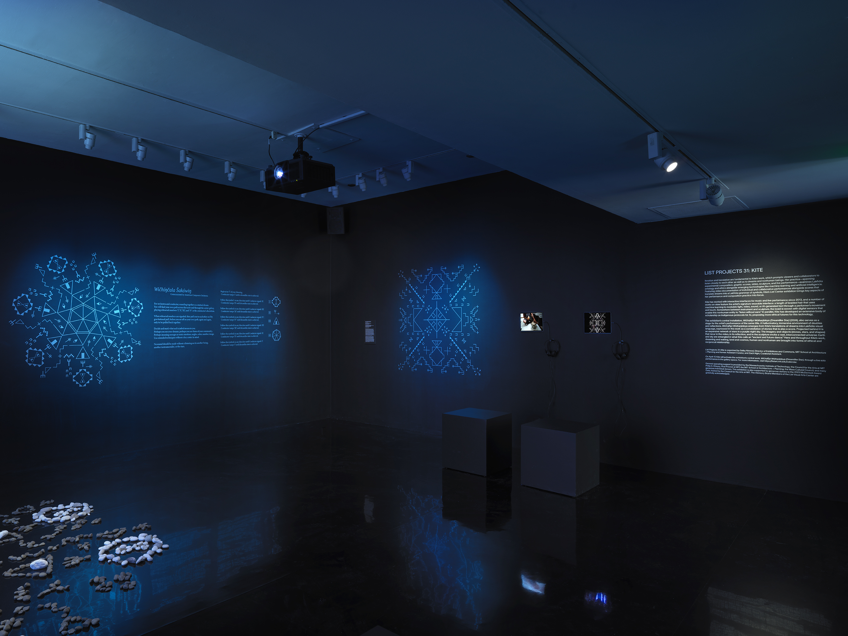 A dimly lit gallery space glows blue. Symbols illuminate the back walls, while‬ ‭ exhibition wall text and iPads displaying video footage are positioned on the far right.‬ ‭ In the lower left corner, a formation of stones peeks into the frame.‬