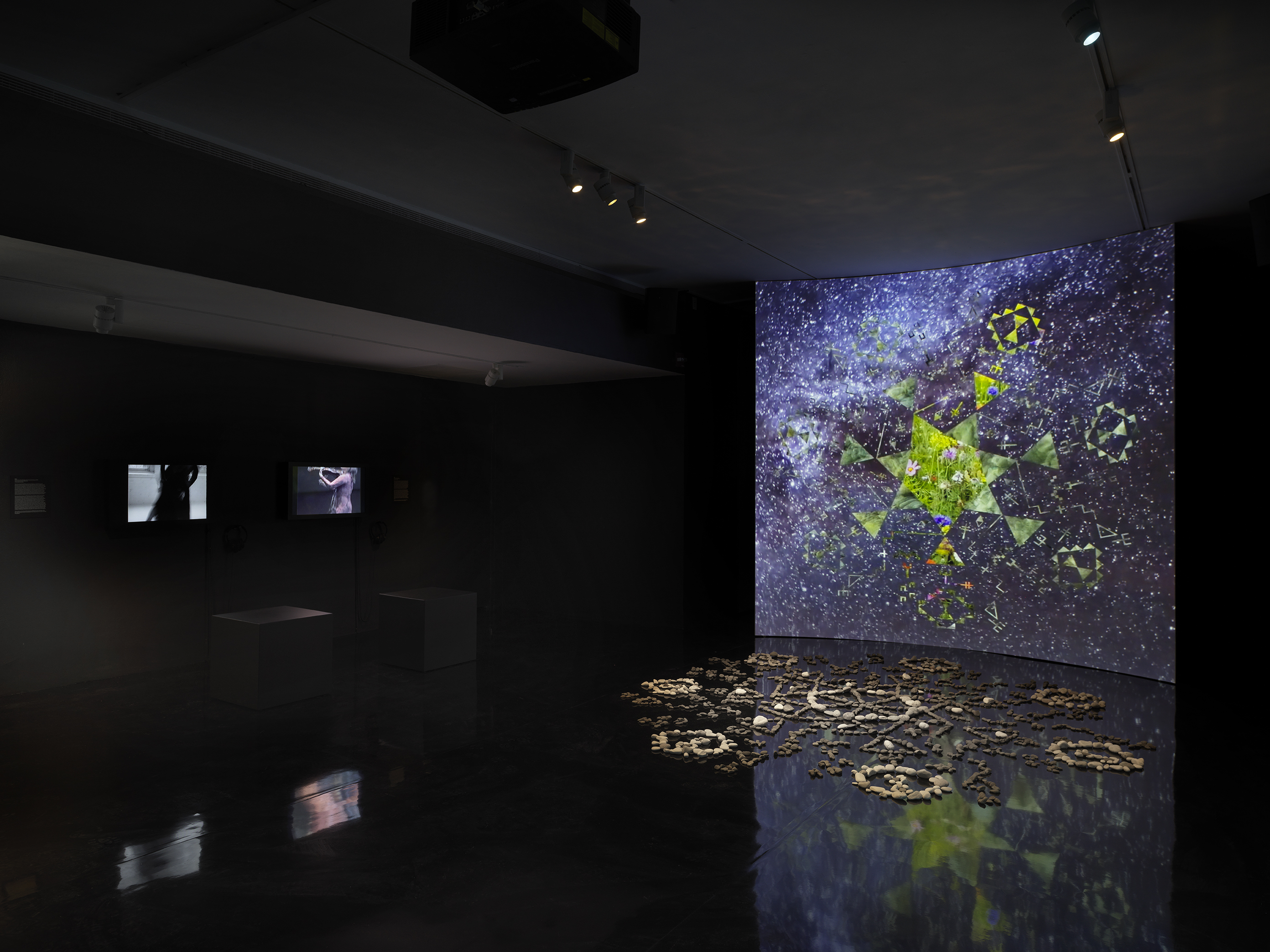 A dark gallery space bathed in tinted blue light. On the right, a large screen‬ ‭ displays a six-sided star at its center, with video footage of green stones projected‬ ‭ within it. Surrounding the star, six inward-facing triangles form a pattern, while‬ ‭ geometric shapes swirl in the background alongside footage of stars and the night‬ ‭ sky. In front of the screen, a formation of stones rests on the reflective black floor.‬ ‭ On the left wall, two videos of performers dancing are displayed.‬