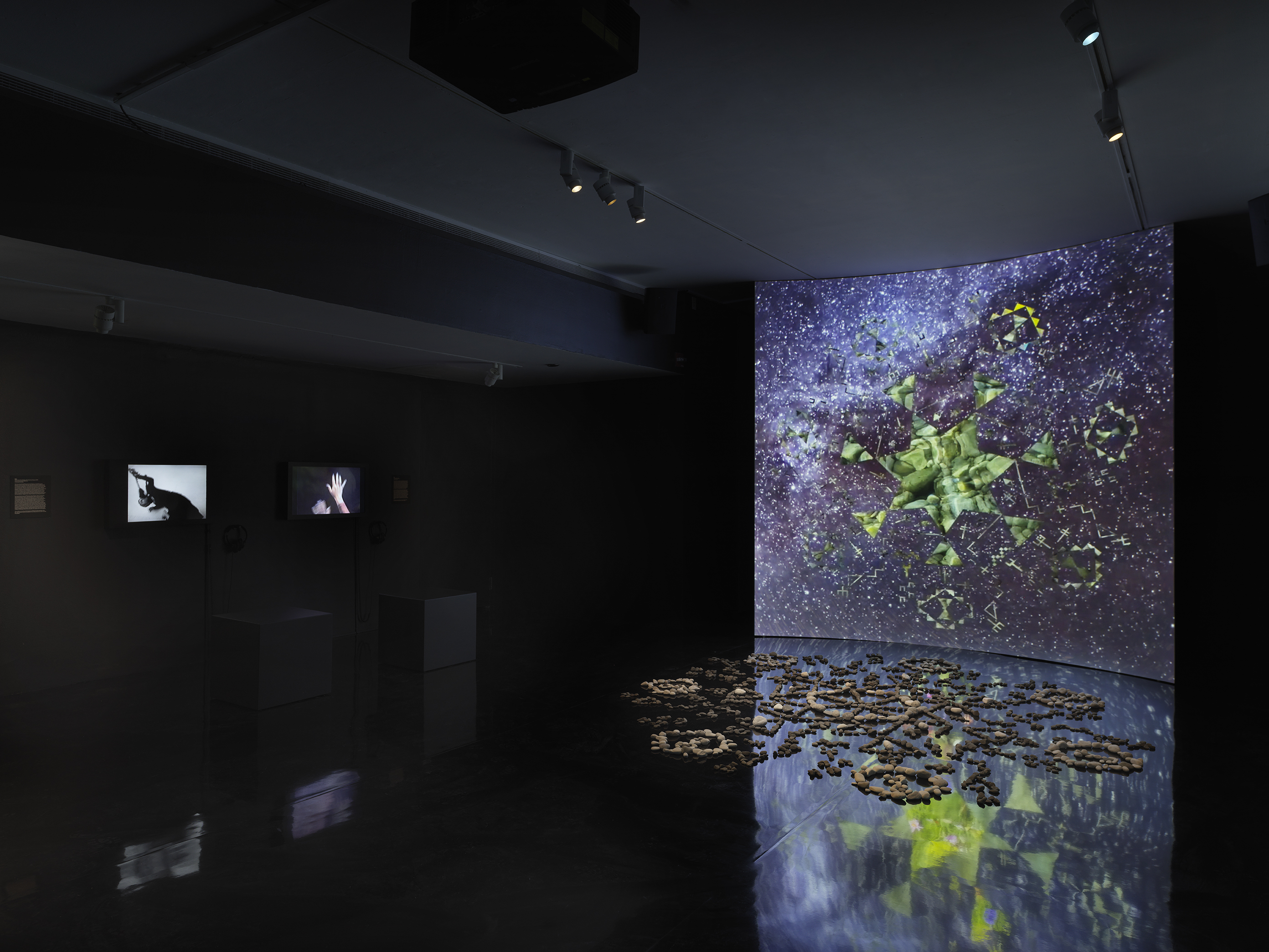 A dark gallery space bathed in tinted blue light. On the right, a large screen‬ ‭ displays a six-sided star at its center, with video footage of green stones projected‬ ‭ within it. Surrounding the star, six inward-facing triangles form a pattern, while‬ ‭ geometric shapes swirl in the background alongside footage of stars and the night‬ ‭ sky. In front of the screen, a formation of stones rests on the reflective black floor.‬ ‭ On the left wall, two videos of performers dancing are displayed.‬