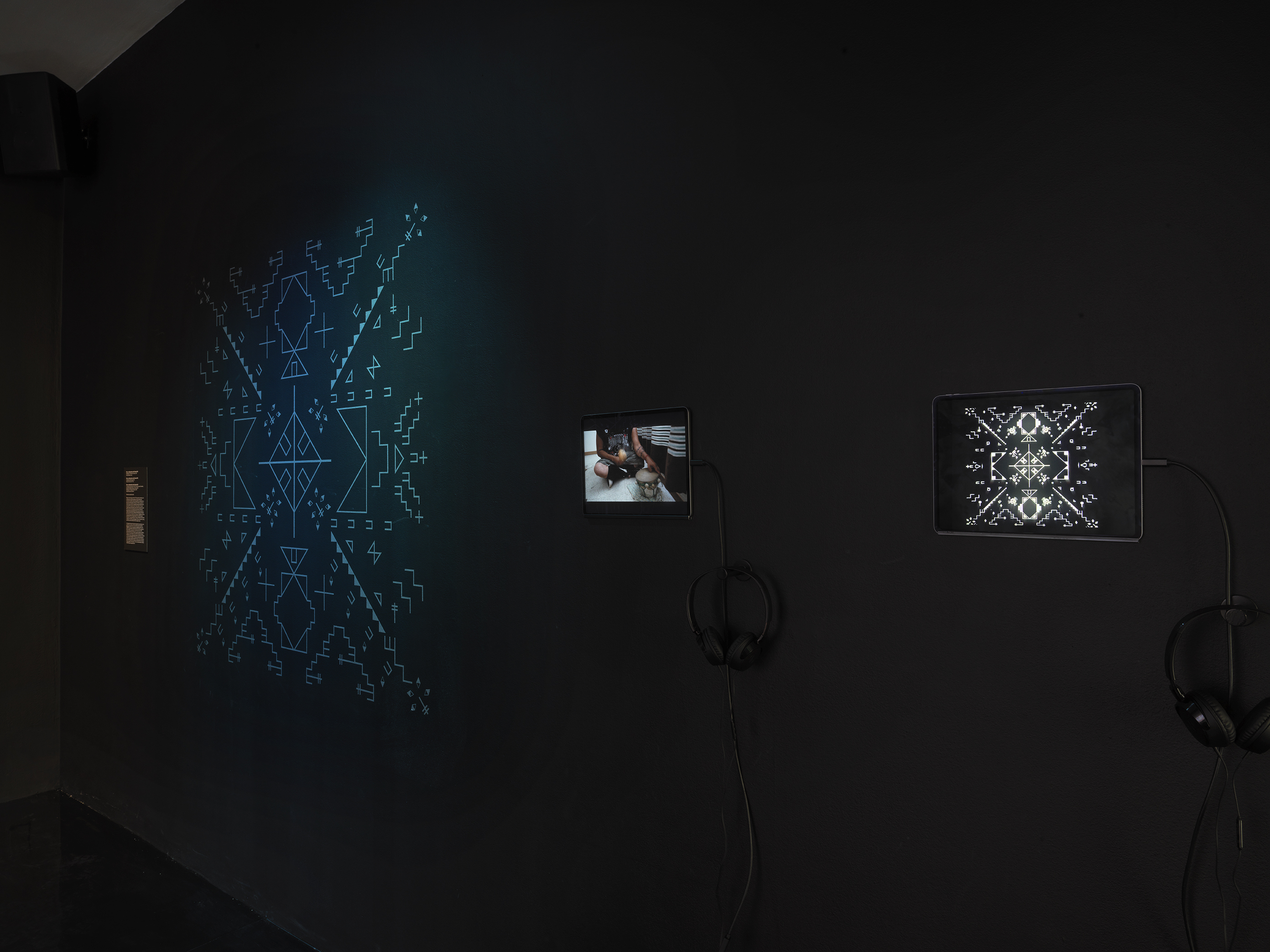 A dimly lit gallery space with two iPads are on display. The far right iPad‬ ‭ shows white geometric forms on a black background, and to its left the other iPad‬ ‭ shows a person drumming. On the far left, a dimly lit form made up of geometric‬ ‭symbols glows blue.‬