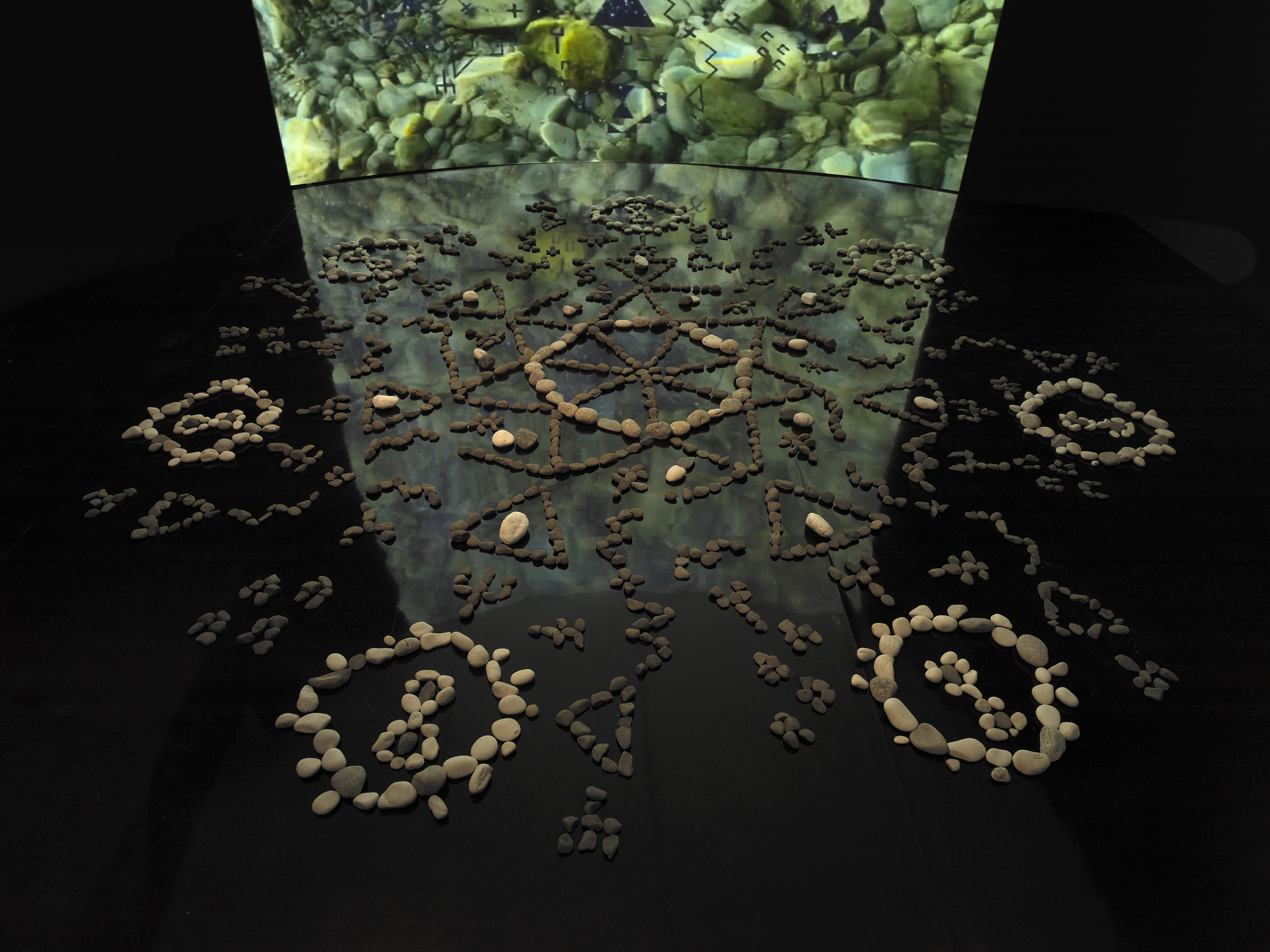 In a dark gallery space, a formation of stones rests on a black mirrored floor.‬ ‭ The stones are placed in different formations, circles, zig-zags, triangles, infinity‬ ‭ symbols, which all meet at a central circular point. Behind the stones is a large‬ ‭ screen showing video footage of rocks under water. The video footage reflects off‬ ‭the mirrored floor.‬‭