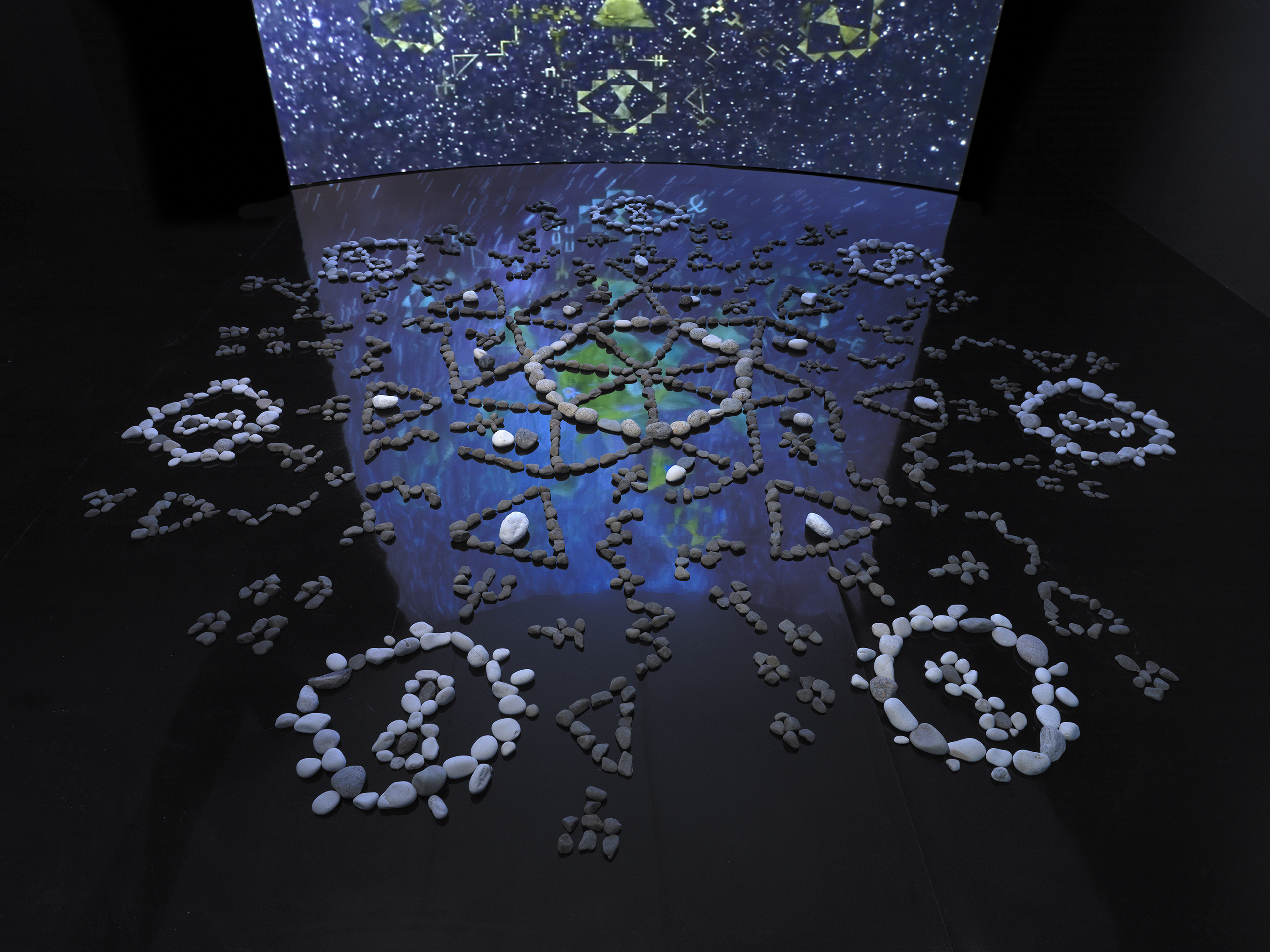 In a dark gallery space, a formation of stones rests on a black mirrored floor.‬ ‭ The stones are placed in different formations, circles, zig-zags, triangles, infinity‬ ‭ symbols, and all meet at a central circular point. Behind the stones is a large screen‬ ‭ showing video footage of stars in the night sky with green geometric forms, similar to‬ ‭ the clusters of stone formations on the floor, circling the sky. The video footage‬ ‭reflects off the mirrored floor.‬ ‭