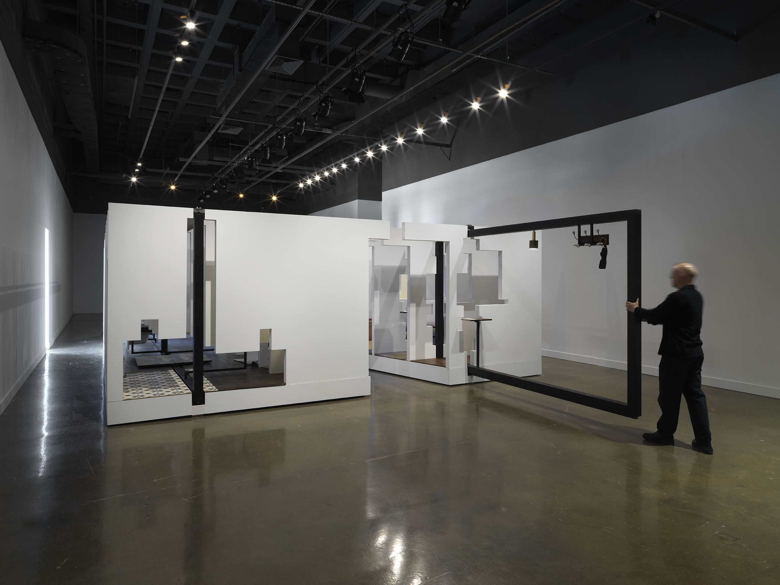 In the middle of an open space, there is a large structure with a series of fragmented white walls divides a living space with wooden floors, furniture, and house plants throughout. Each section of wall is connected by black beams. A man in a all black pulls one of the black beams on the right side of the frame.