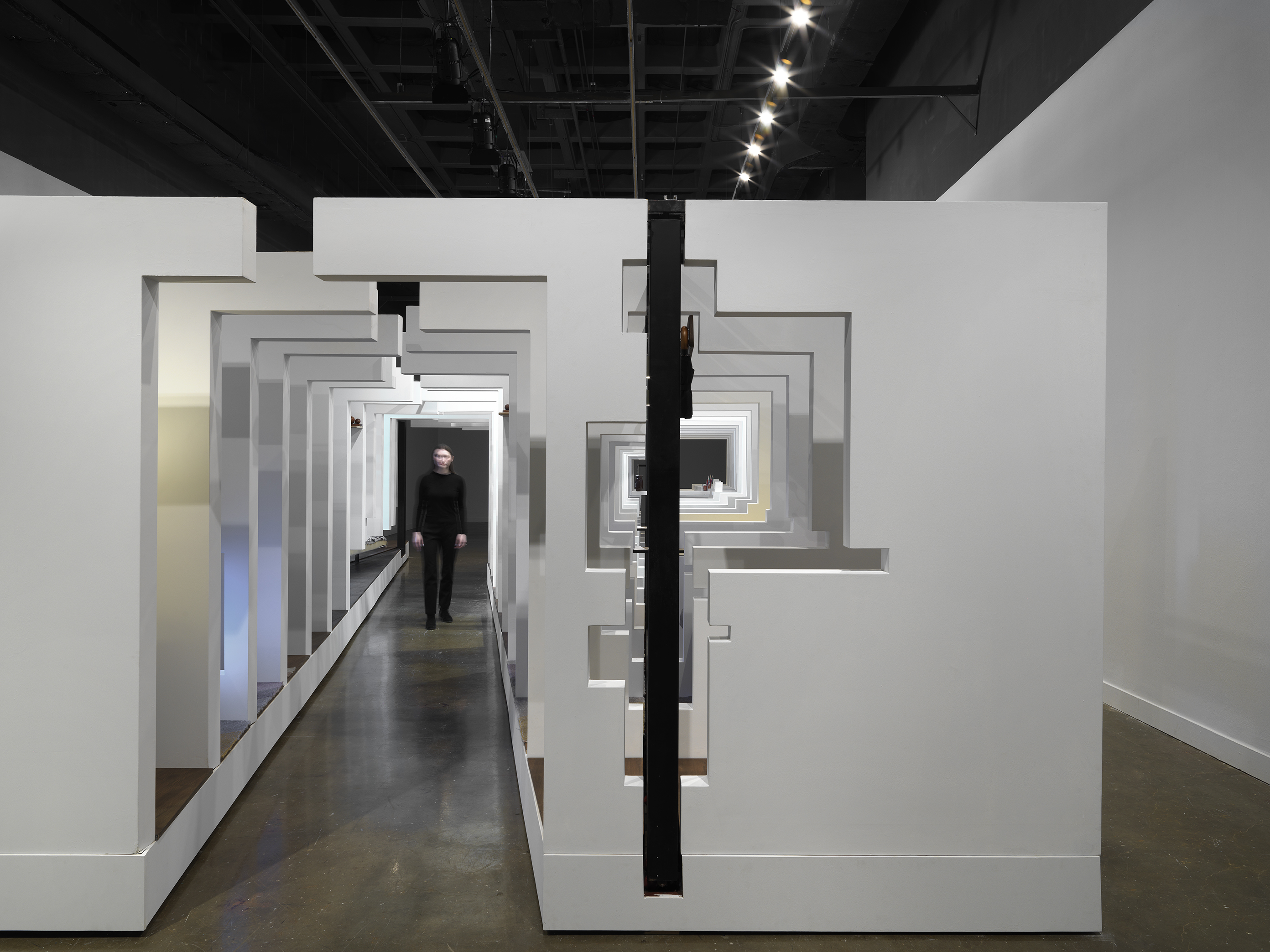 A series of fragmented white walls divides a living space with wooden floors, furniture, and house plants throughout. Each section of wall is connected by black beams. A woman in a all black walks through an opening in the middle of the structure.
