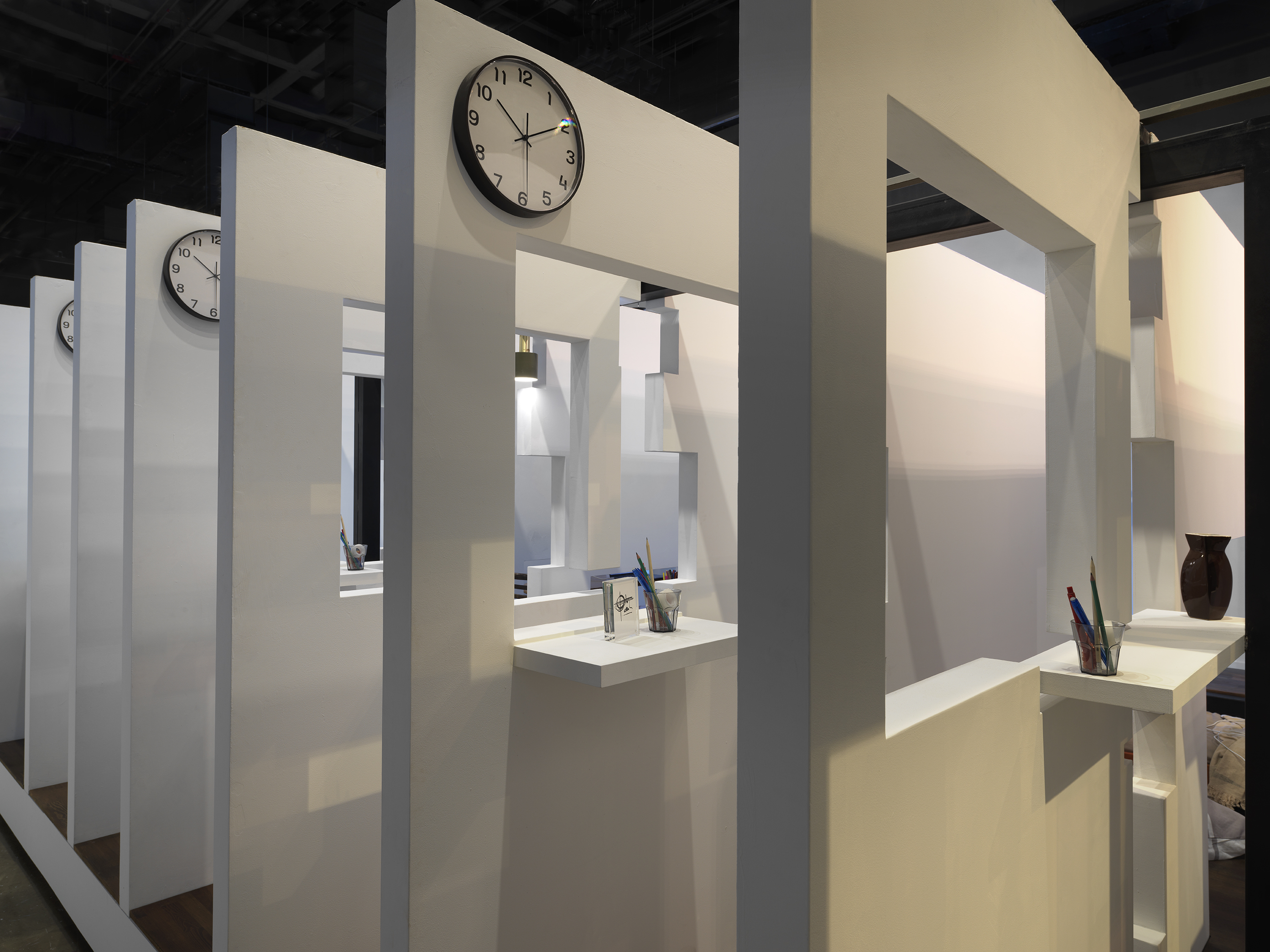 A series of white walls with a window cutout has a view of the next white wall with a window cutout. There is an analog clock hanging at the top left corner on every wall.