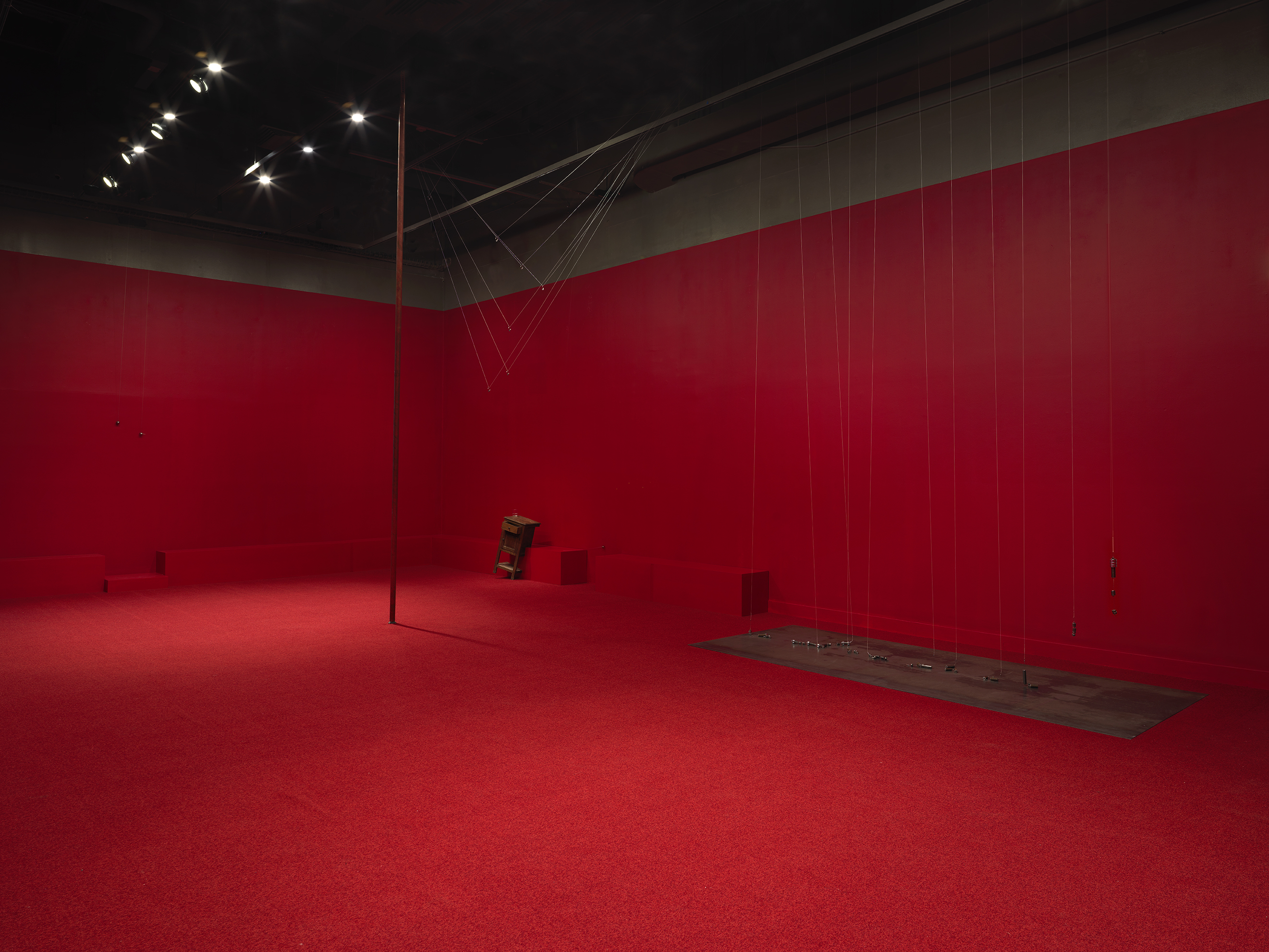 A copper poll stands vertical in the middle of a completely red painted walled room with red carpeting. To the right side there are a series of weights on strings above a metal plate. A wooden cabinet tilted on a angle is visible against the wall behind. Three metal spheres hang on strings suspended from the ceiling throughout the room.
