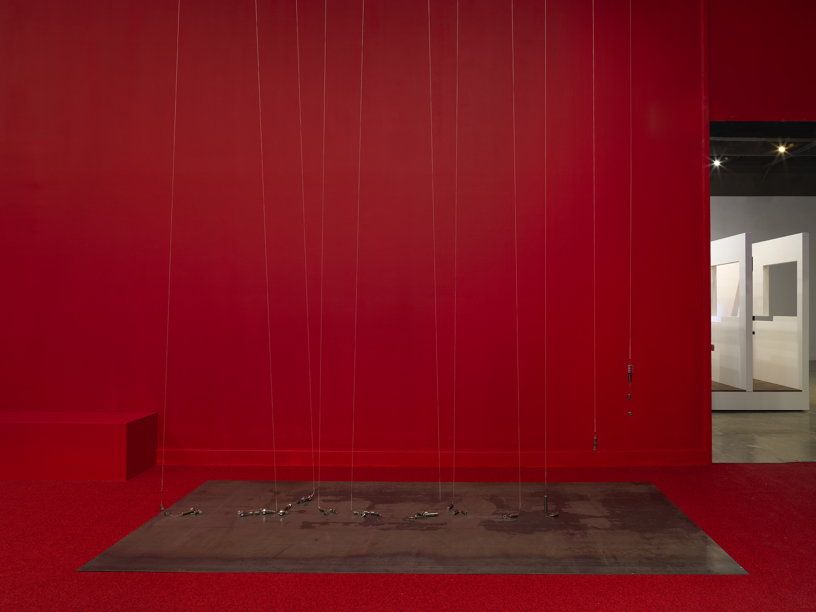A series of weights suspended on strings above a metal plate in the middle of a completely red painted walled room with red carpeting. On the right side there is a doorway to bright room.