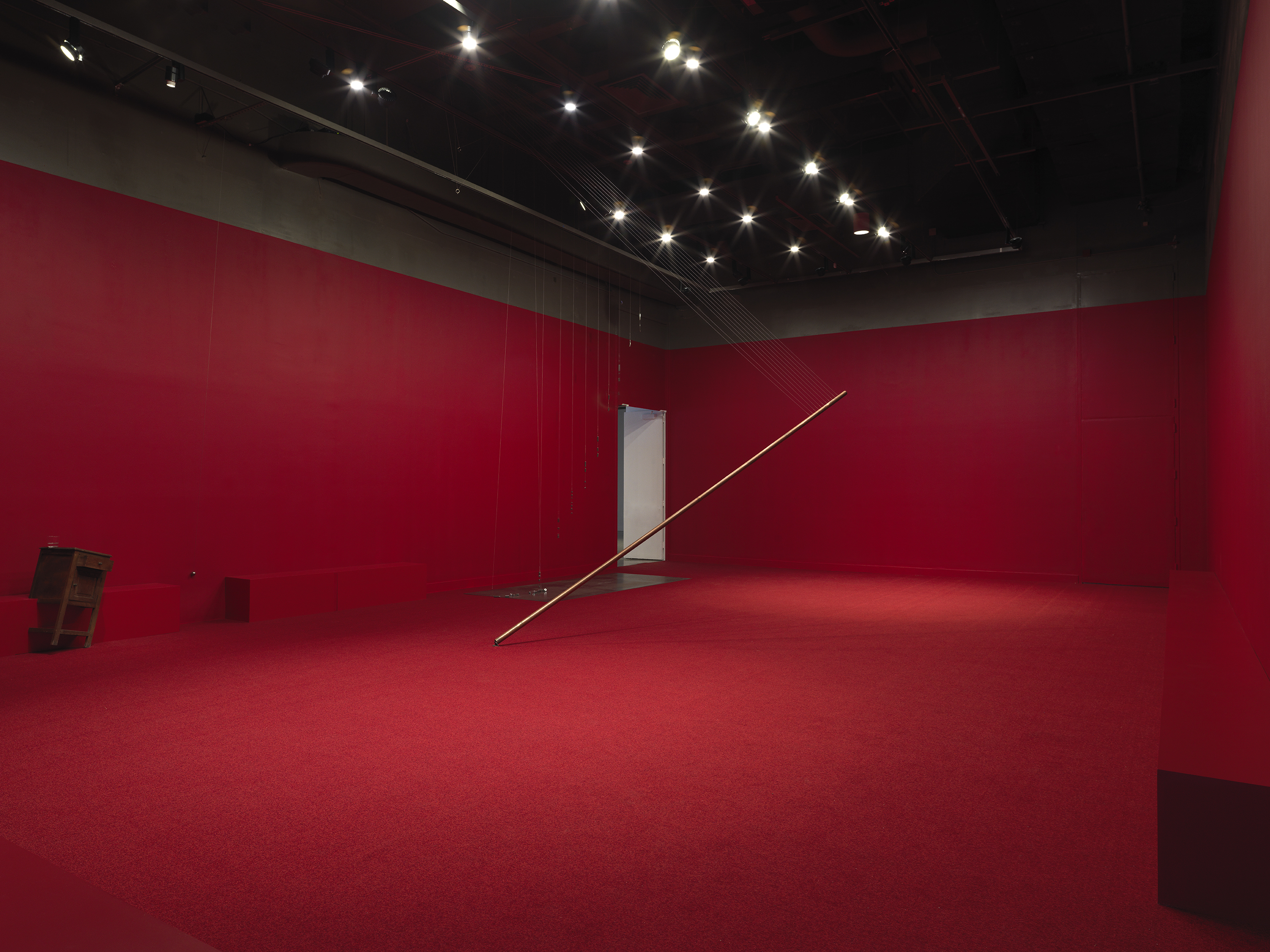 A copper poll suspended on an angle in the middle of a completely red painted walled room with red carpeting. Behind there are a series of weights on strings above a metal plate. A wooden cabinet tilted on a angle is visible against the wall to the left of the frame. Metal spheres hang on strings suspended from the ceiling throughout the room.