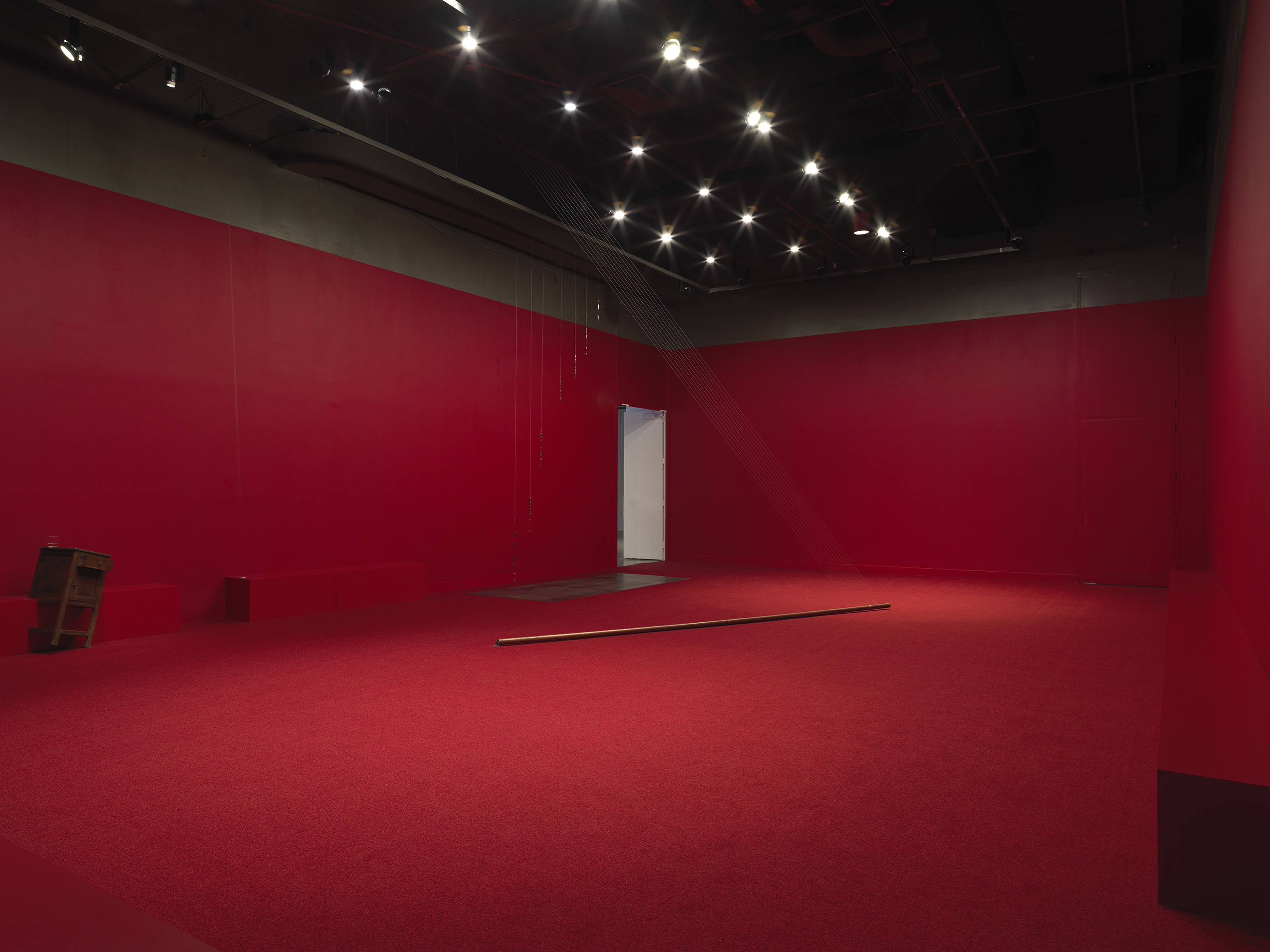 A copper poll suspended on an angle in the middle of a completely red painted walled room with red carpeting. Behind there are a series of weights on strings above a metal plate. A wooden cabinet tilted on a angle is visible against the wall to the left of the frame. Metal spheres hang on strings suspended from the ceiling throughout the room.