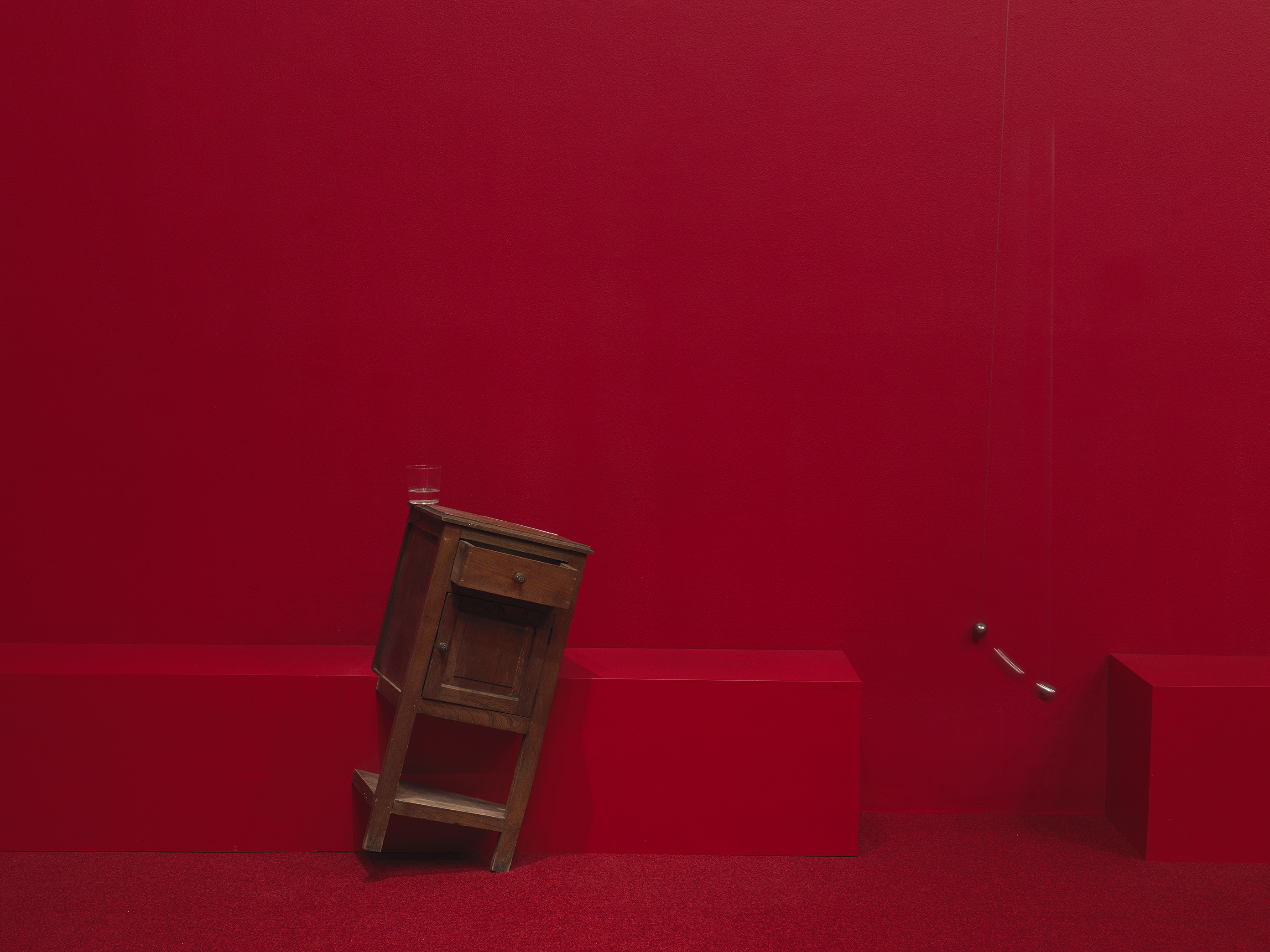 A wooden cabinet tilted on a angle to the right in a completely red painted room with red carpeting. A swinging metal sphere on a string suspended from the ceiling is captured in motion.