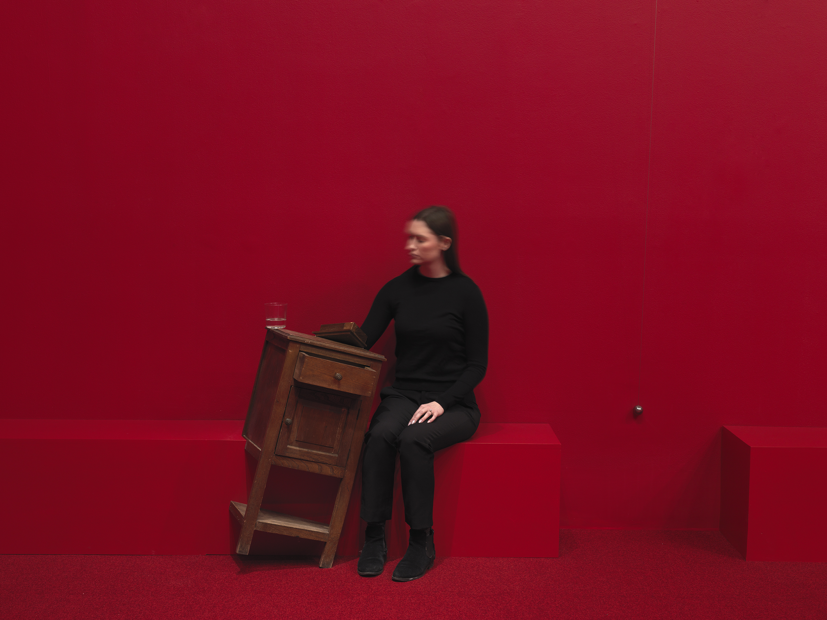 A wooden cabinet tilted on a angle to the right in a completely red painted room with red carpeting. A woman in a black sits to the right with her hand inside of the cabinet. A swinging metal sphere on a string suspended from the ceiling is captured in motion.
