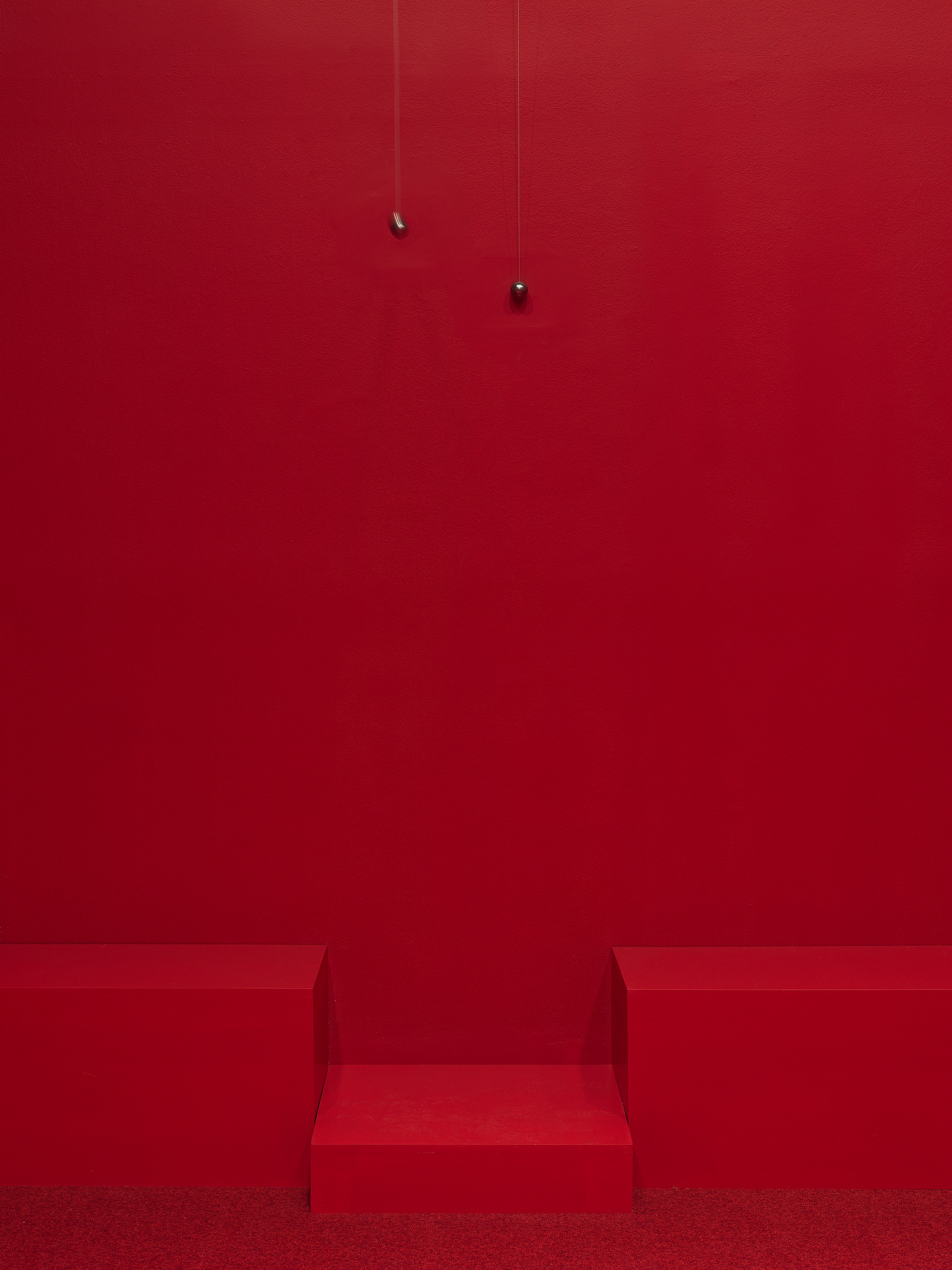 Two swinging metal spheres suspended from the ceiling on strings are captured in motion in a completely red painted walled room with red carpet.