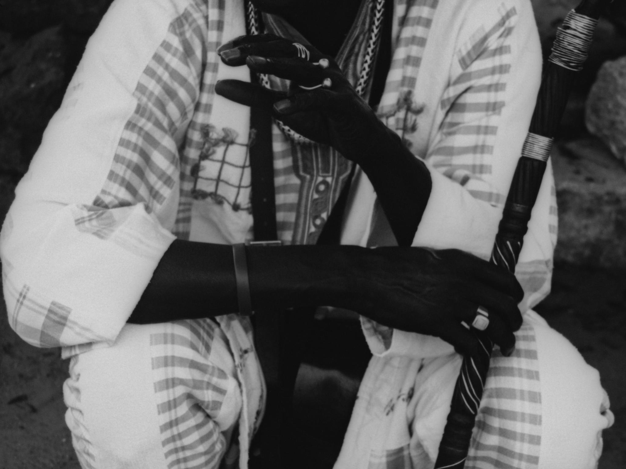 Black and white video still of a man sitting down holding a scepter wearing matching patterned clothing.