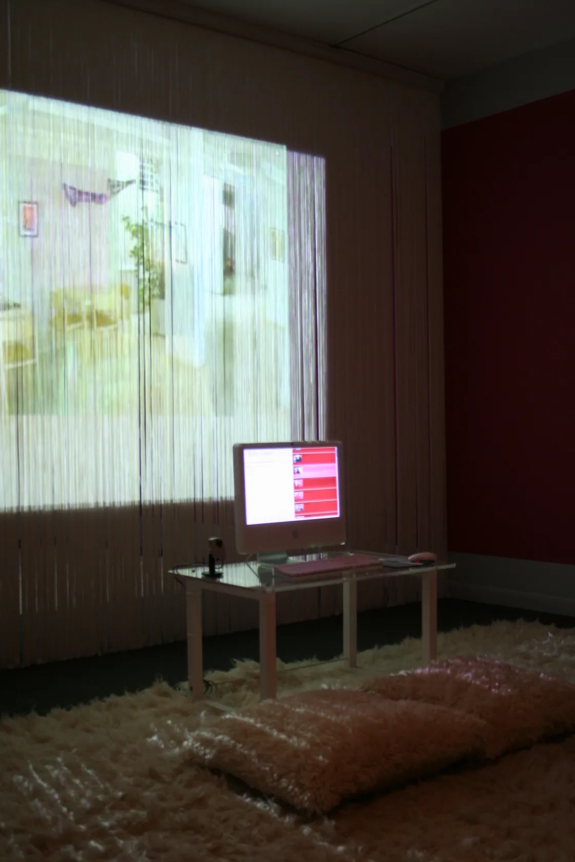 A mac computer monitor sits on a small table close to the ground. Beneath it is a plush rug and pillow.