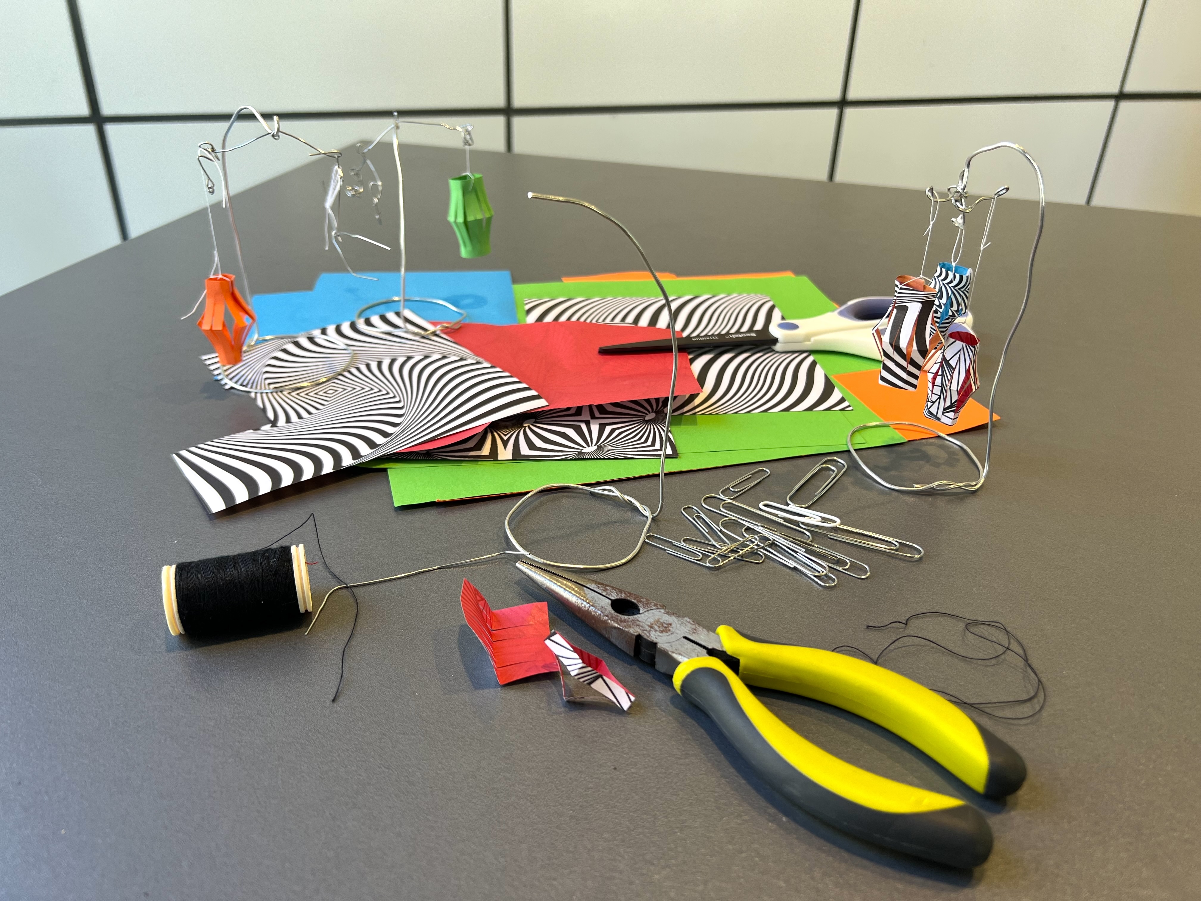 There are three small wire mobile's with a green, orange and multicolor paper lanterns on a gray table. There is a group of paper clips, a stack of unfolded paper, and a pair of bright yellow wire cutters in the foreground.