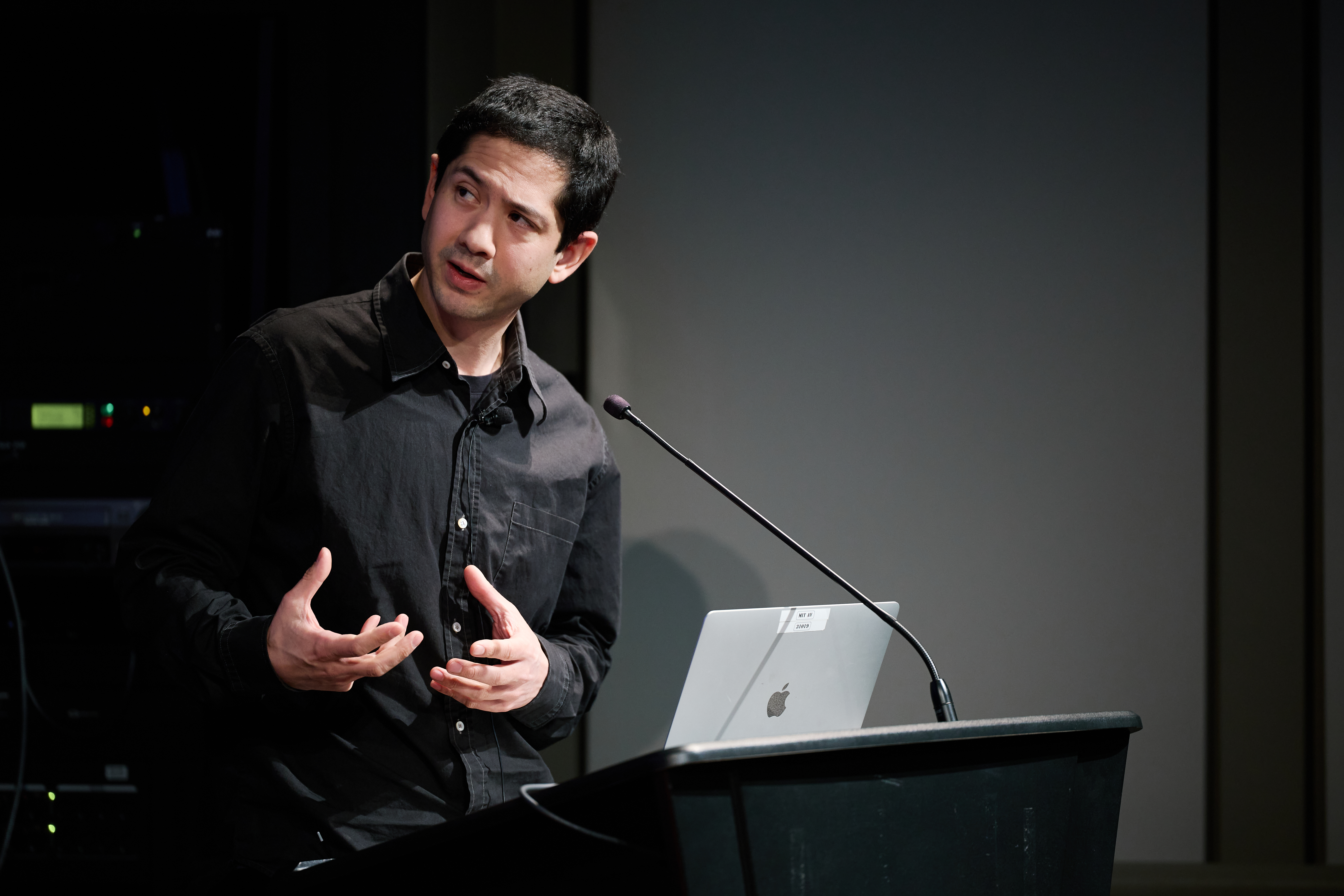A man speaking and looking to the upper left corner of the frame while standing behind a podium with a tall microphone and a silver Mac laptop. The background is dark behind except for green lights from the sound system.