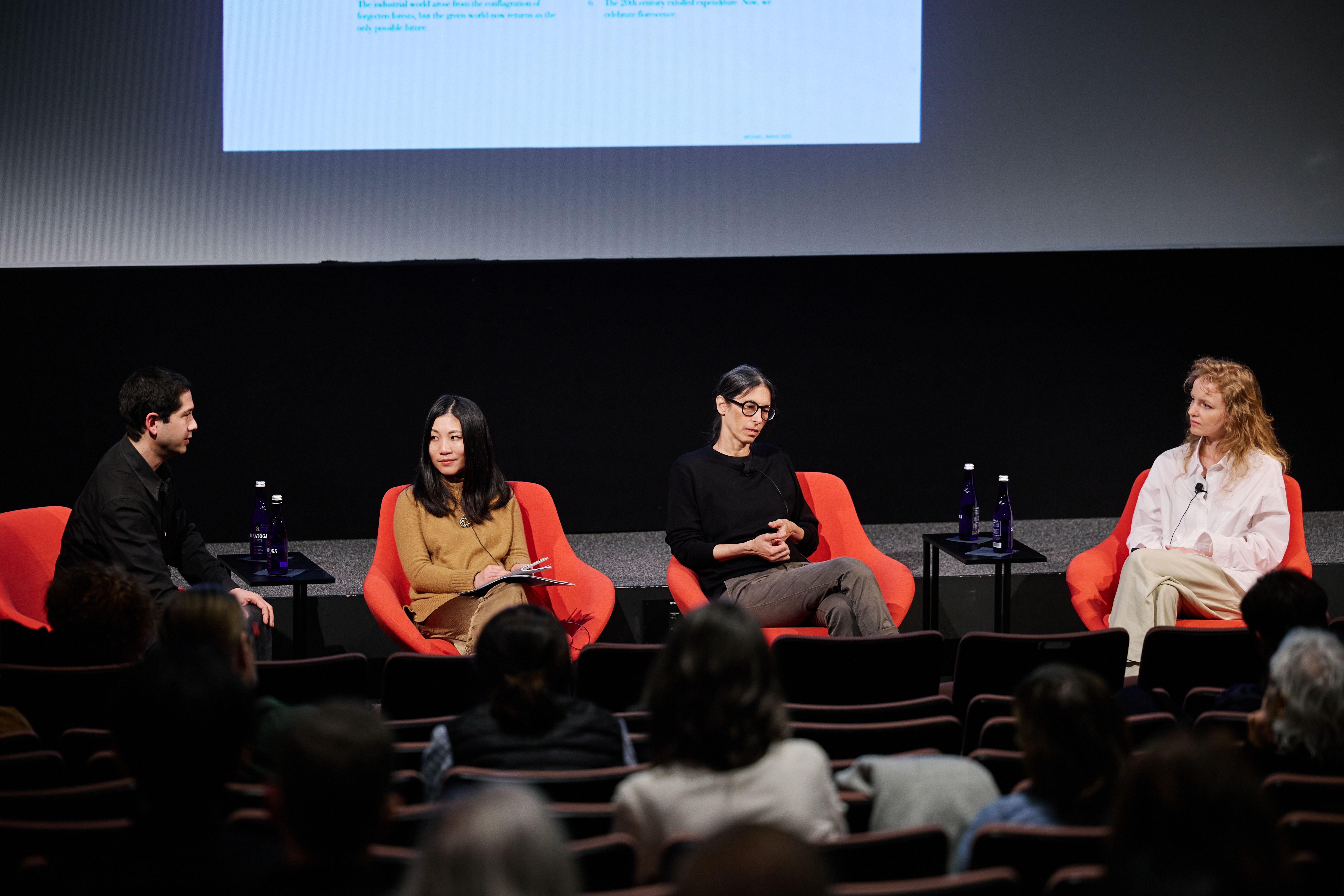 Four people sitting in a line in bright red chairs on a stage in front of a projector screen. The person on the far left side of the frame is speaking to the three other individuals.