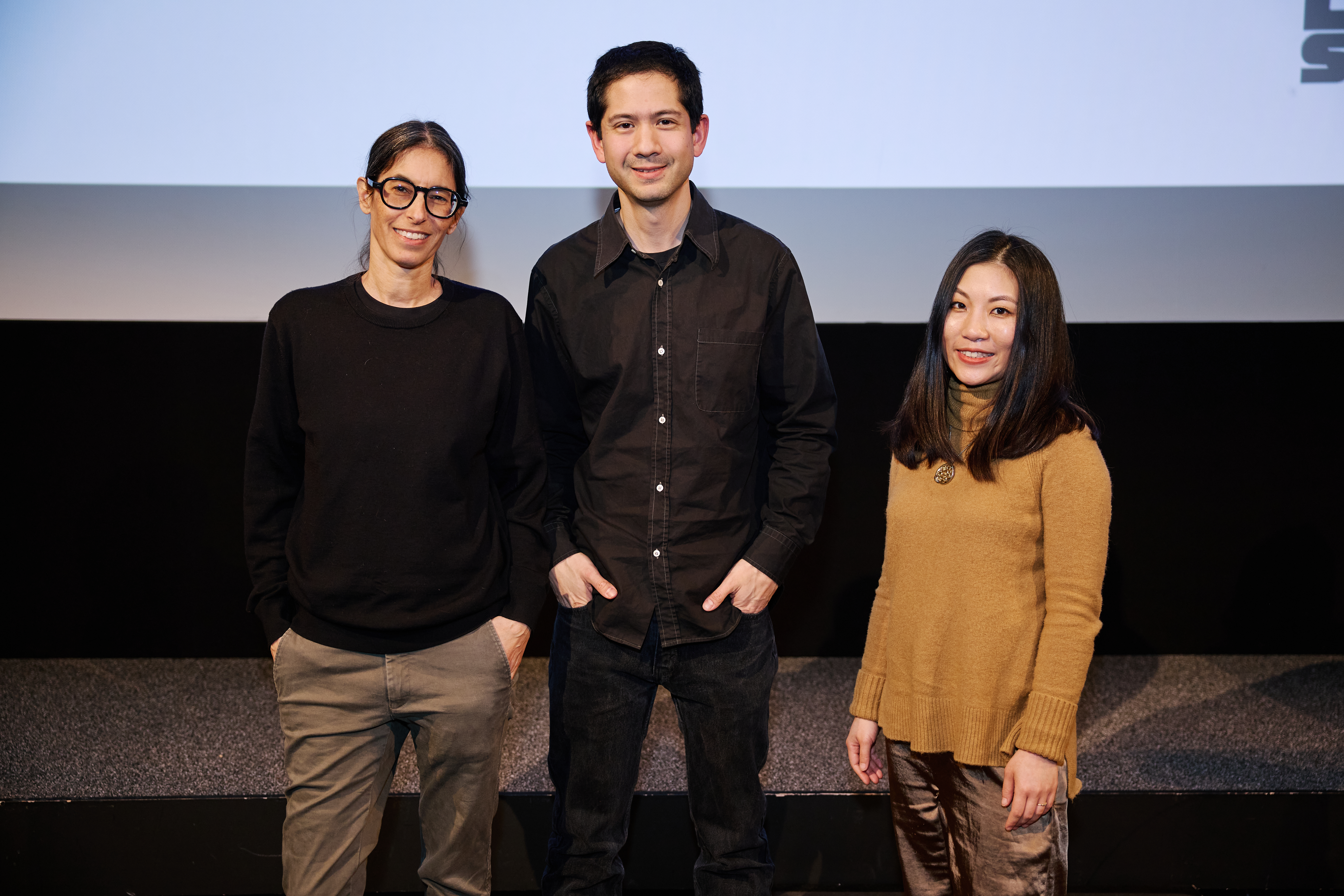 Three people standing on a stage in front of a projector screen. The person in the middle is taller than the people on either side.