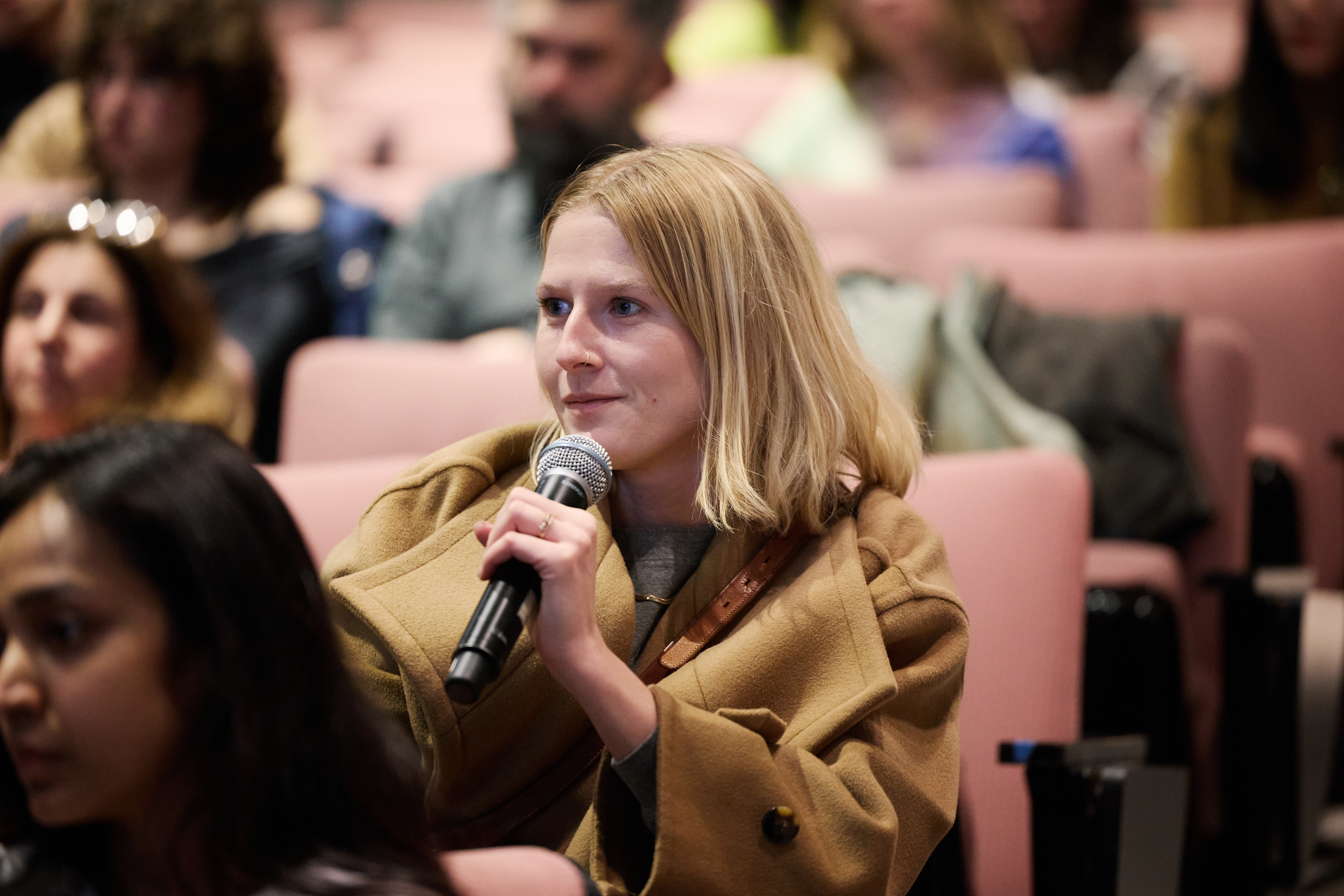 Woman with short blonde hair in a camel colored jacket sitting in a theater audience, holding a microphone near her mouth.