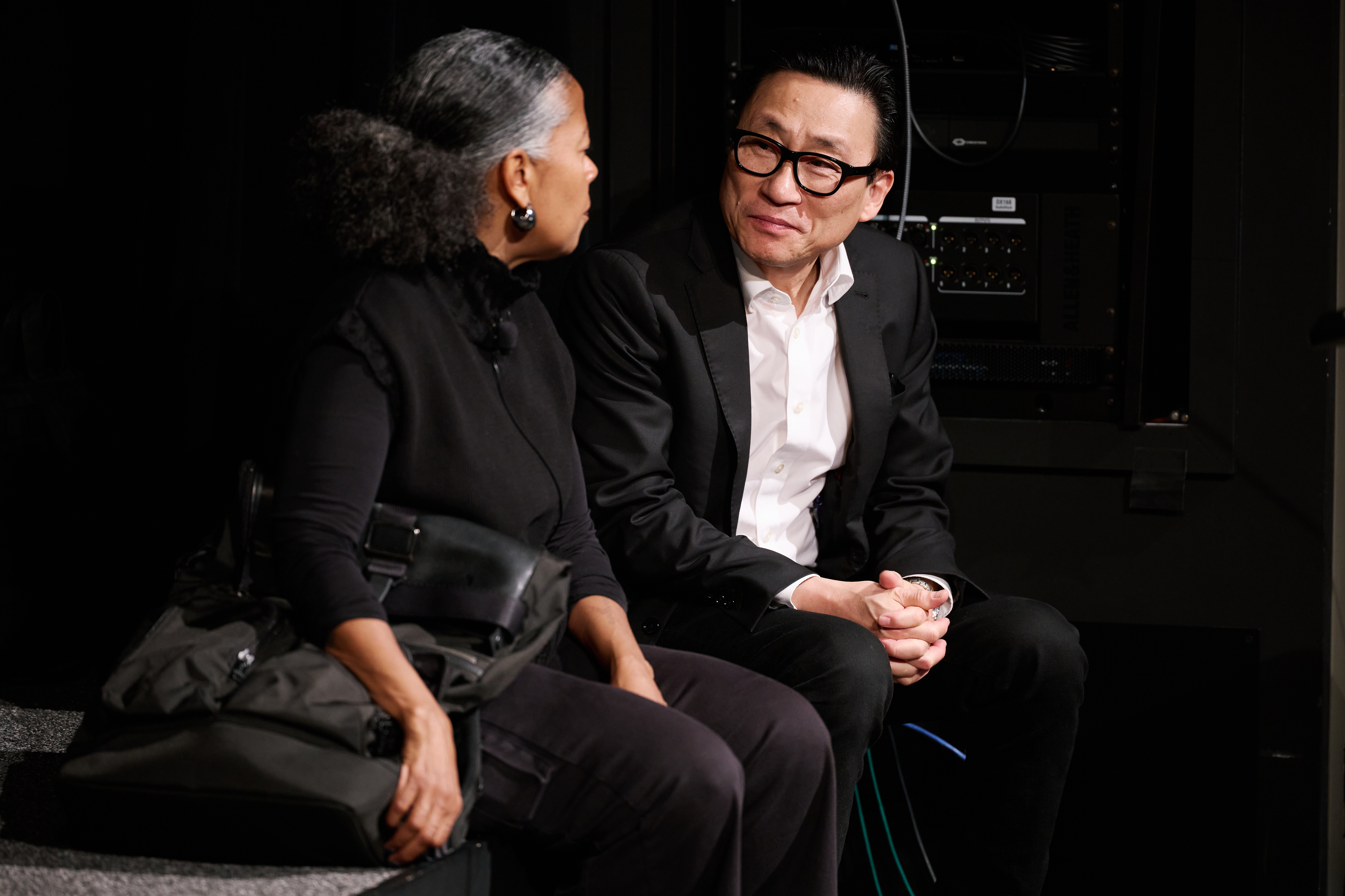 A man and woman sitting on a stage. The background is dark and the figures are illuminated by stage light.