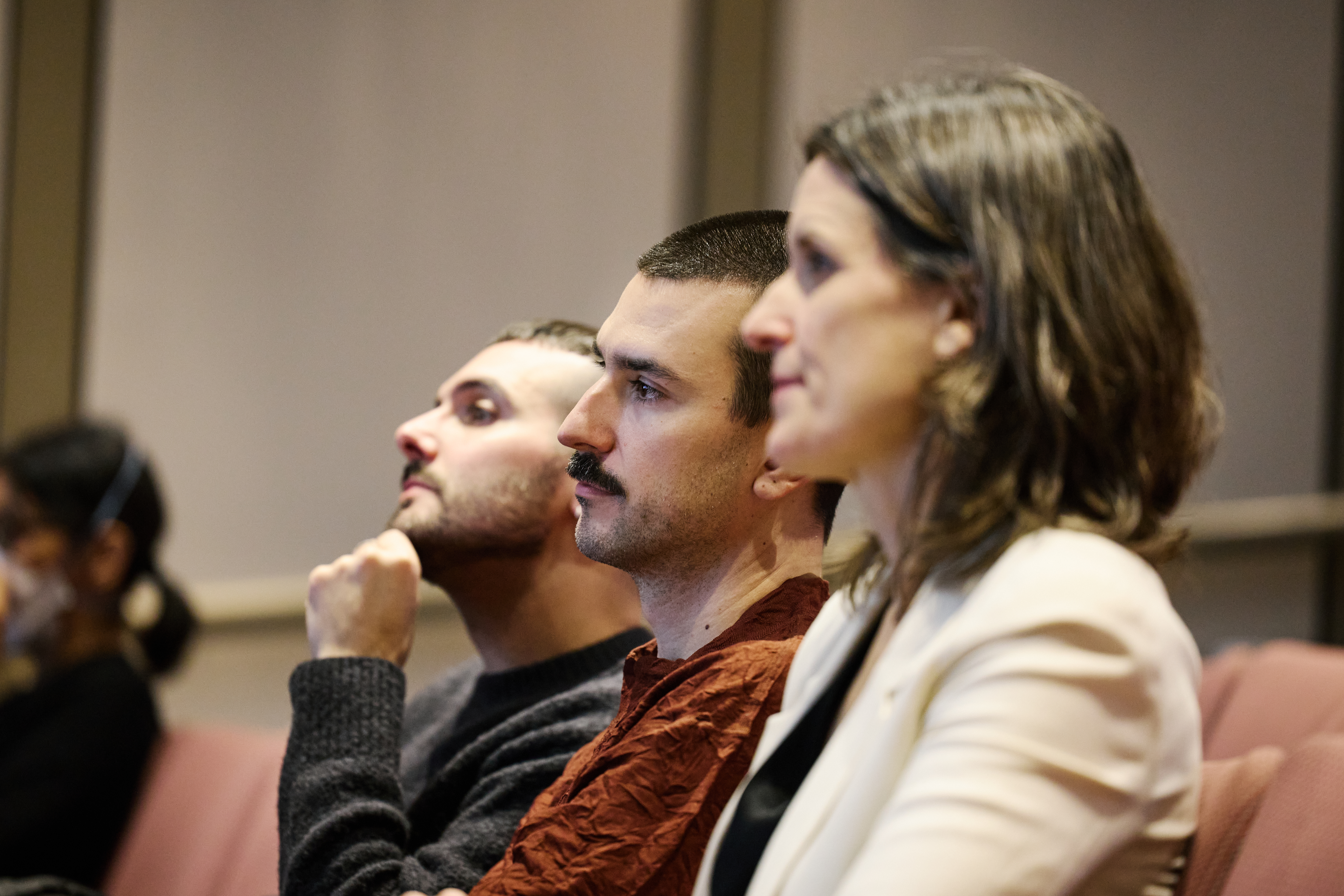 Three people sitting in red theater chairs face the left side of the image, looking at something out of the frame. The person furthest from the camera has their hand on their chin inquisitively.