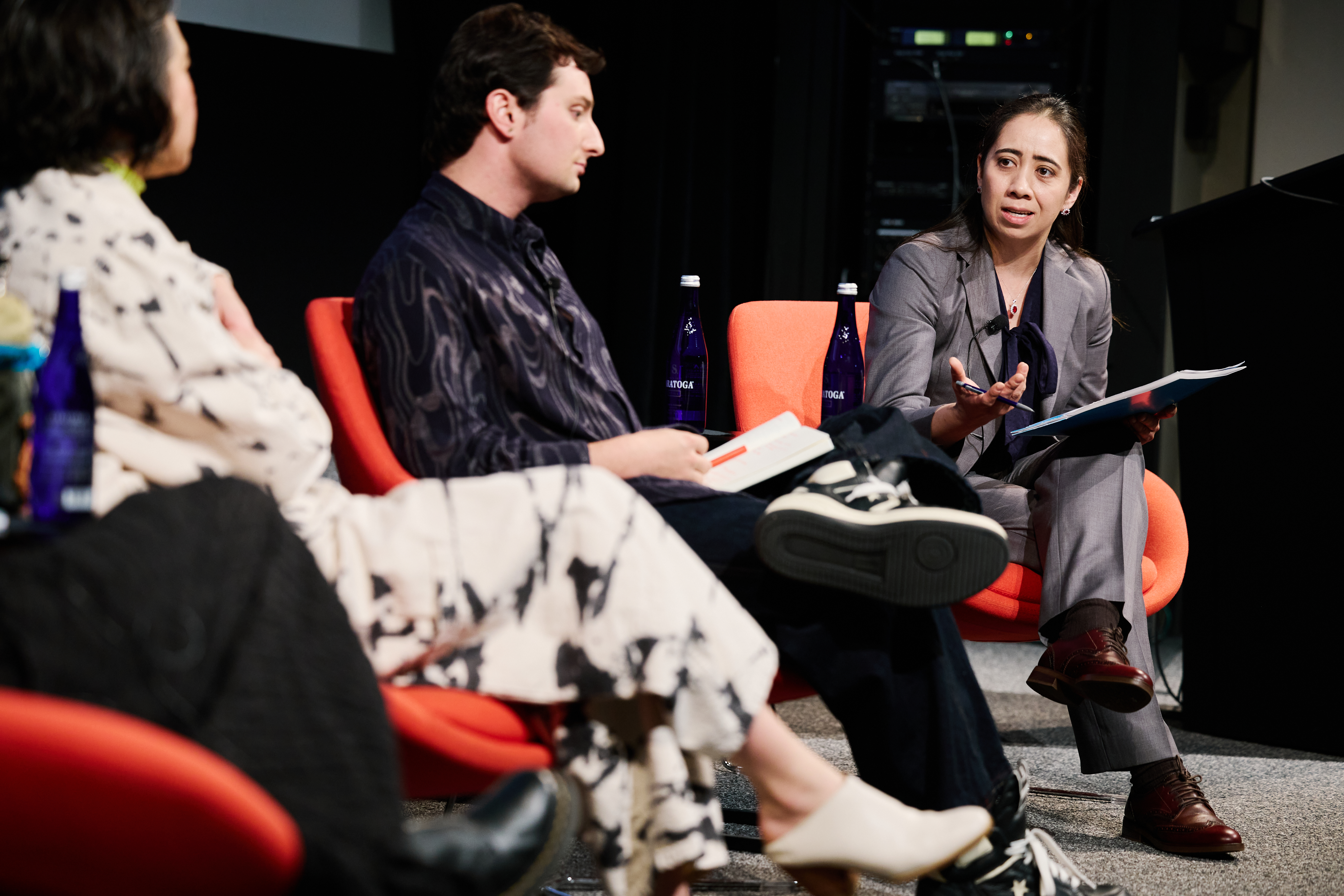 Three people sitting in a line in bright red chairs on a stage. The person furthest from the camera is speaking to the panel.