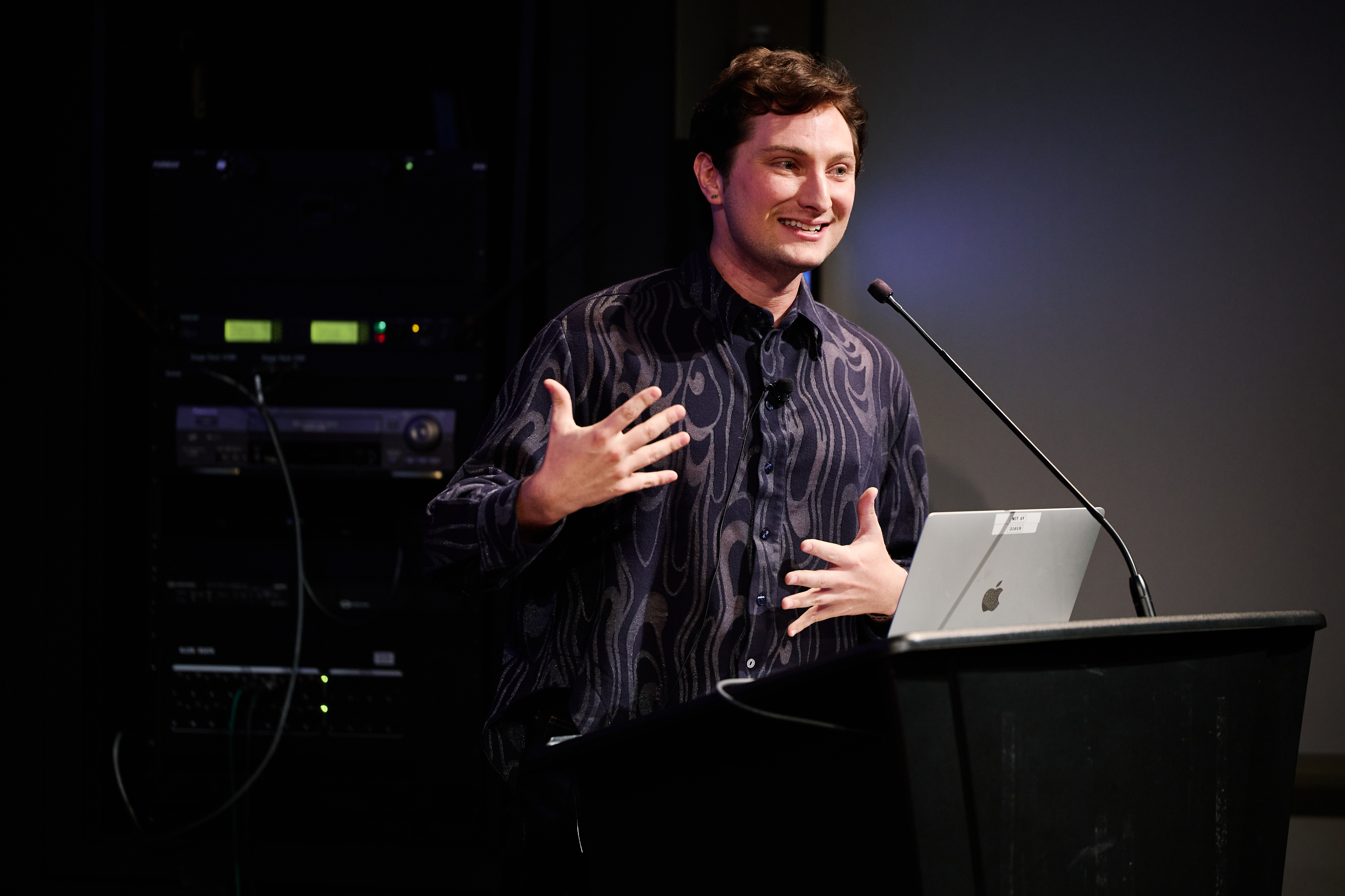 Person in a navy buttoned long sleeve shirt is talking with their hands in front of their chest, standing behind a podium with a tall microphone and a silver Mac laptop.