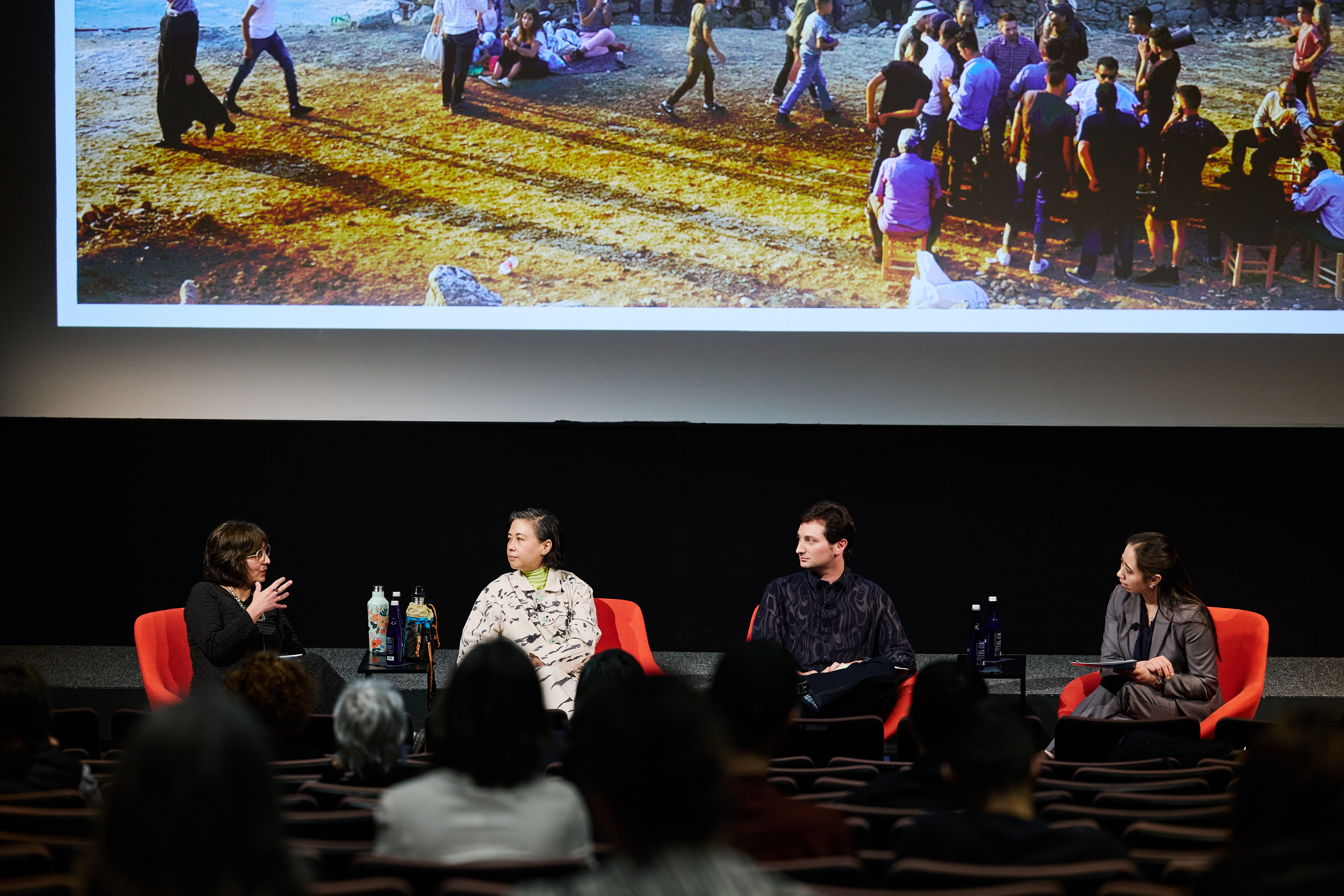 Four people sitting in a line in bright red chairs on a stage in front of a projector screen. The person on the furthest left hand side of the stage is speaking to the panelist.