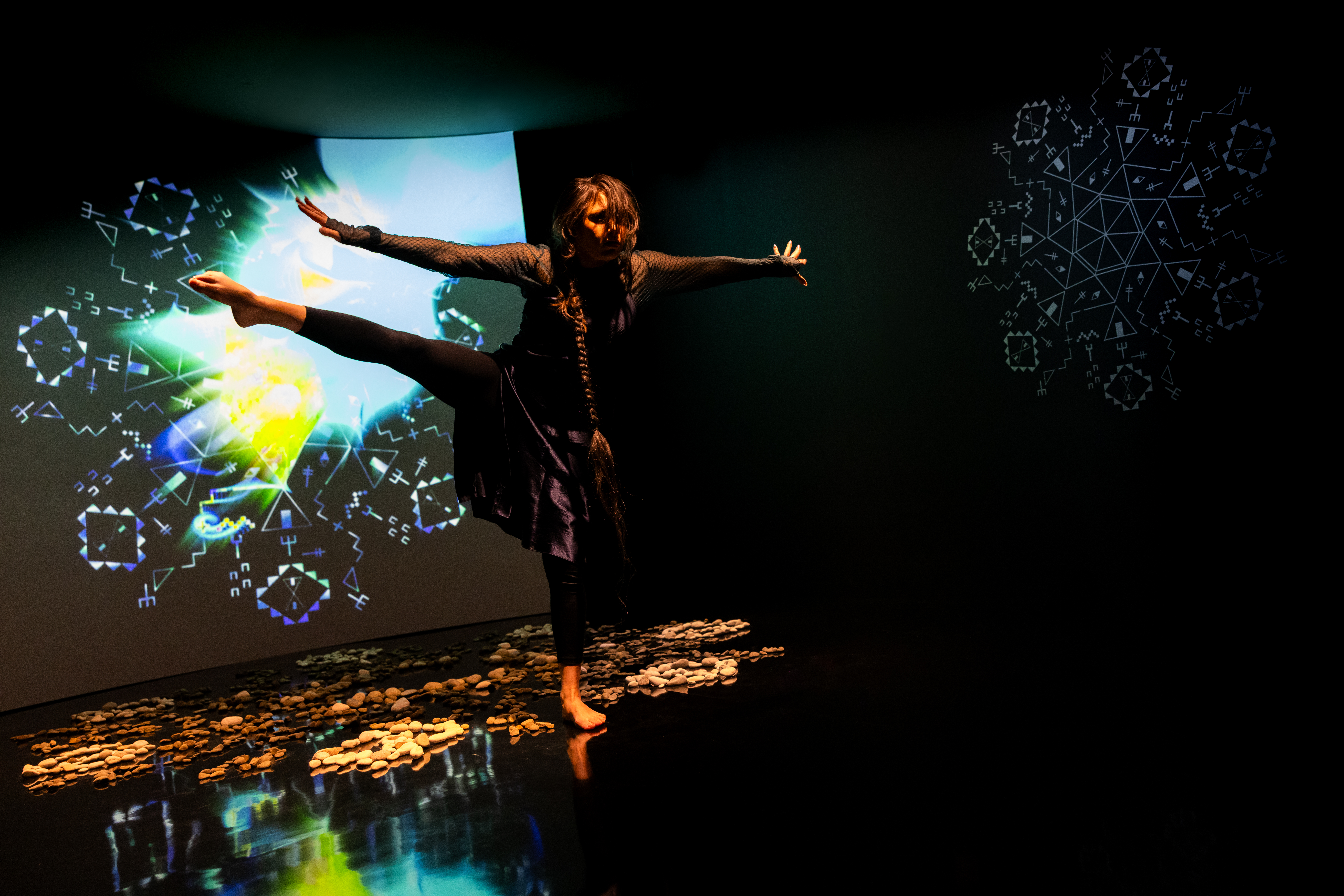Still of a performance showcasing a woman spotlighted on stage with her arms out to both sides facing the camera and her right leg raised to hip height with foot pointed. A brightly colored geometric pattern in the background is juxtaposed by a black wall.