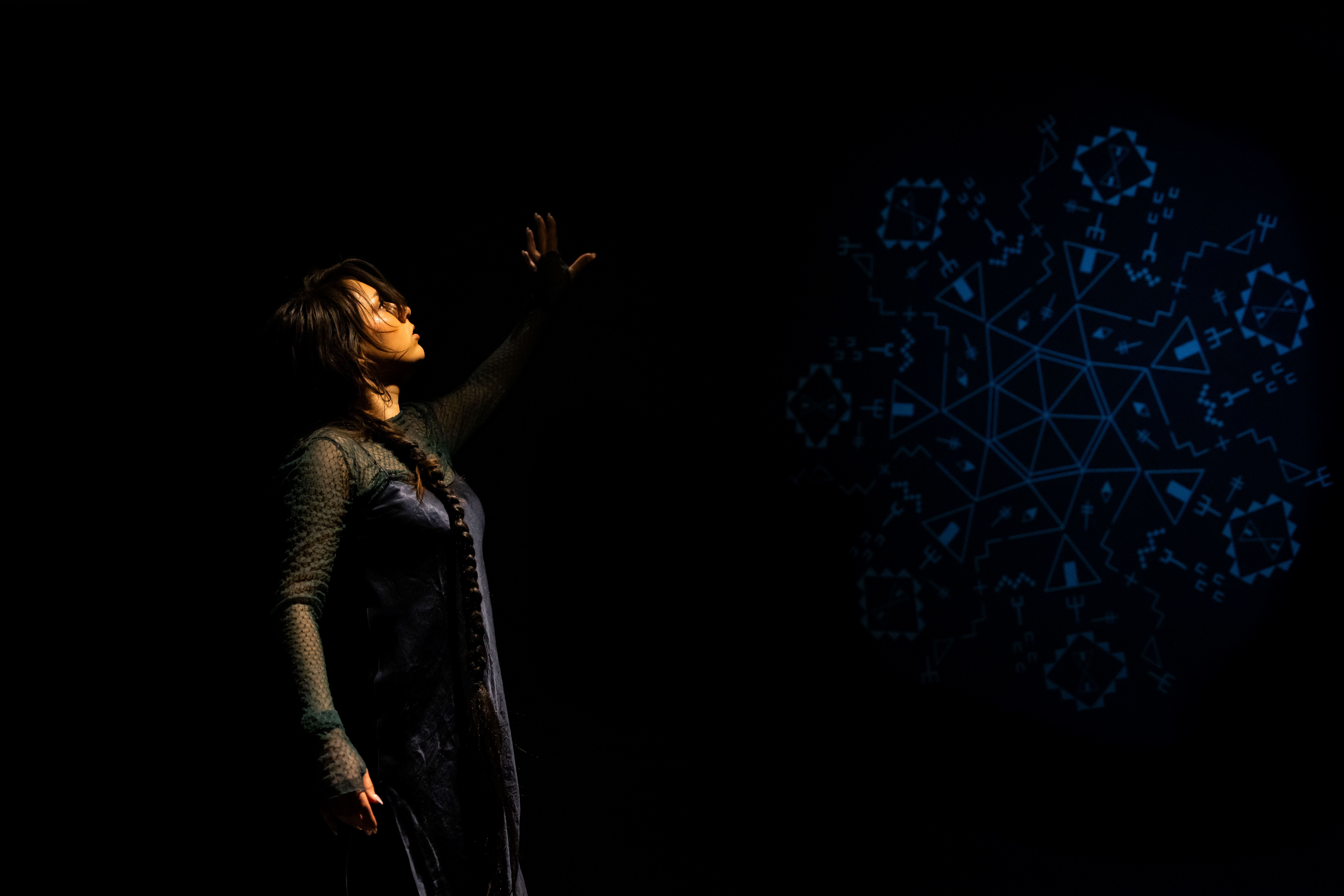 Still of a performance showcasing a woman spotlighted on stage with her left arm raised above her head. A faint colored geometric pattern in the background is juxtaposed by a black wall.