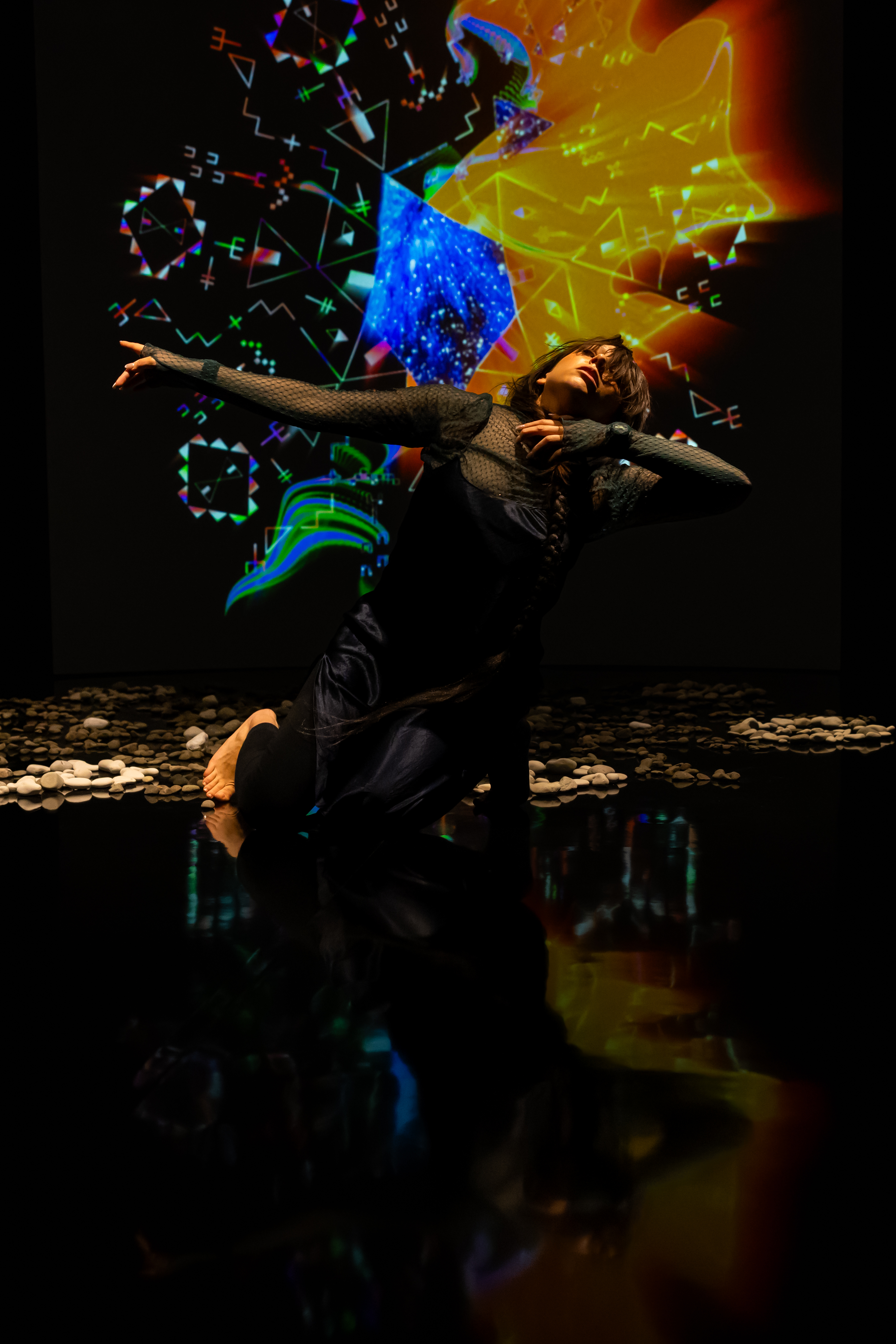 Still of a performance showcasing a woman spotlighted on stage with her arms raised out to the right side of her body facing the camera. A brightly colored geometric pattern in the background is juxtaposed by a black wall.