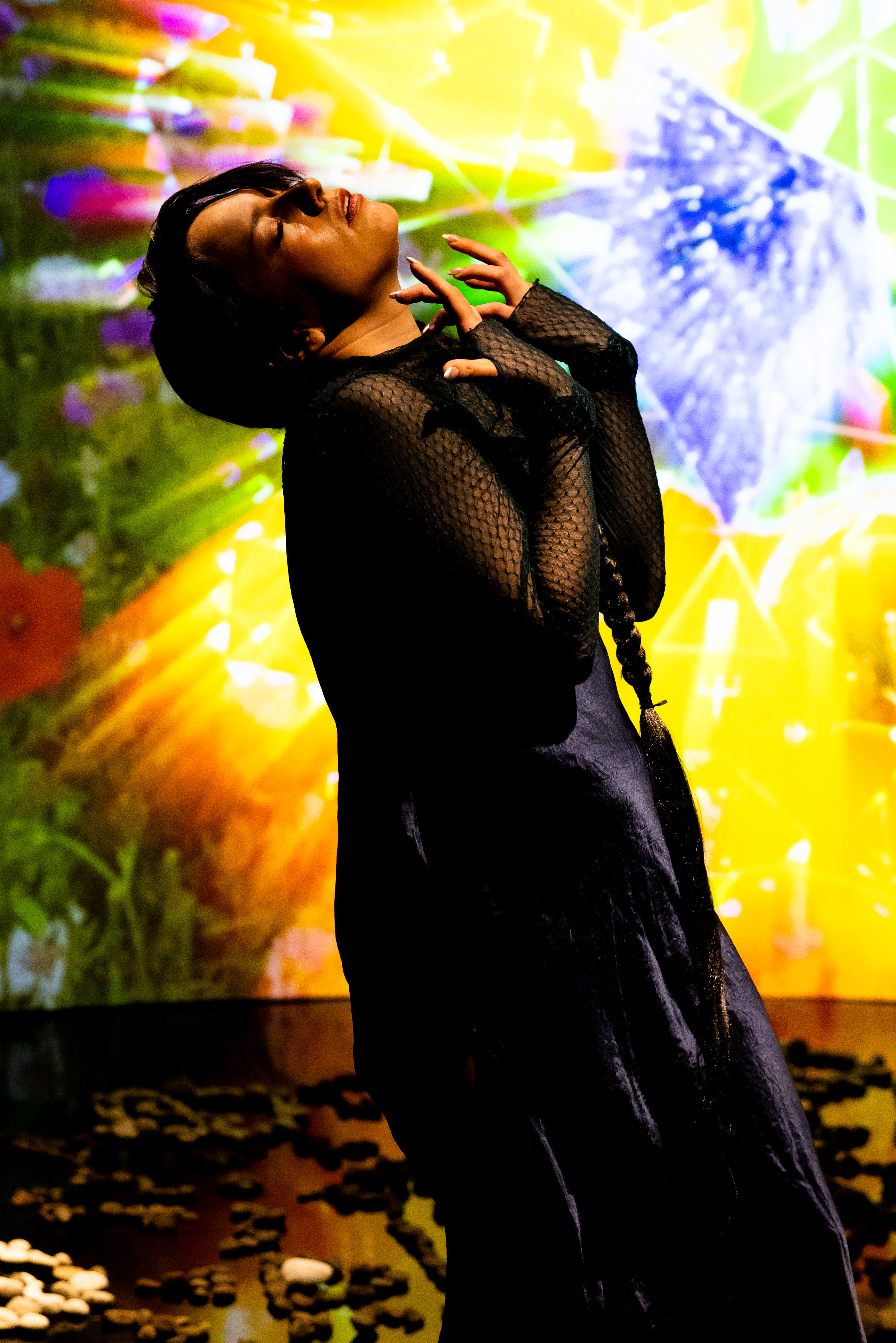Still of a performance showcasing a woman spotlighted on stage with her head tilted up, arms raised in front of her chest. A brightly colored geometric pattern in the background is juxtaposed by a black mirrored floor.