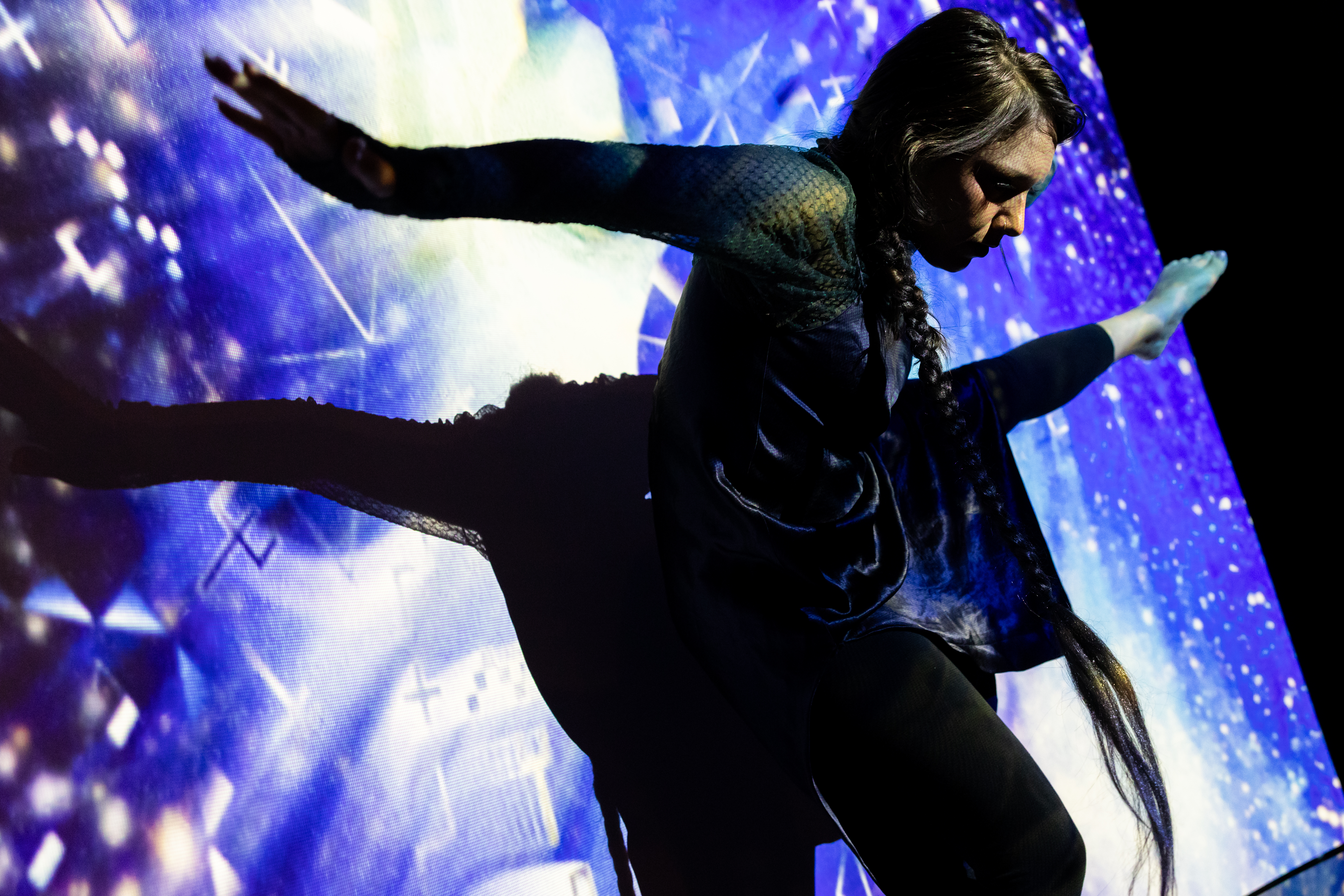 Still of a performance showcasing a woman facing the camera spotlighted on stage with her arms out to both sides and her right leg raised to hip height with foot pointed. A brightly colored geometric pattern in the background.