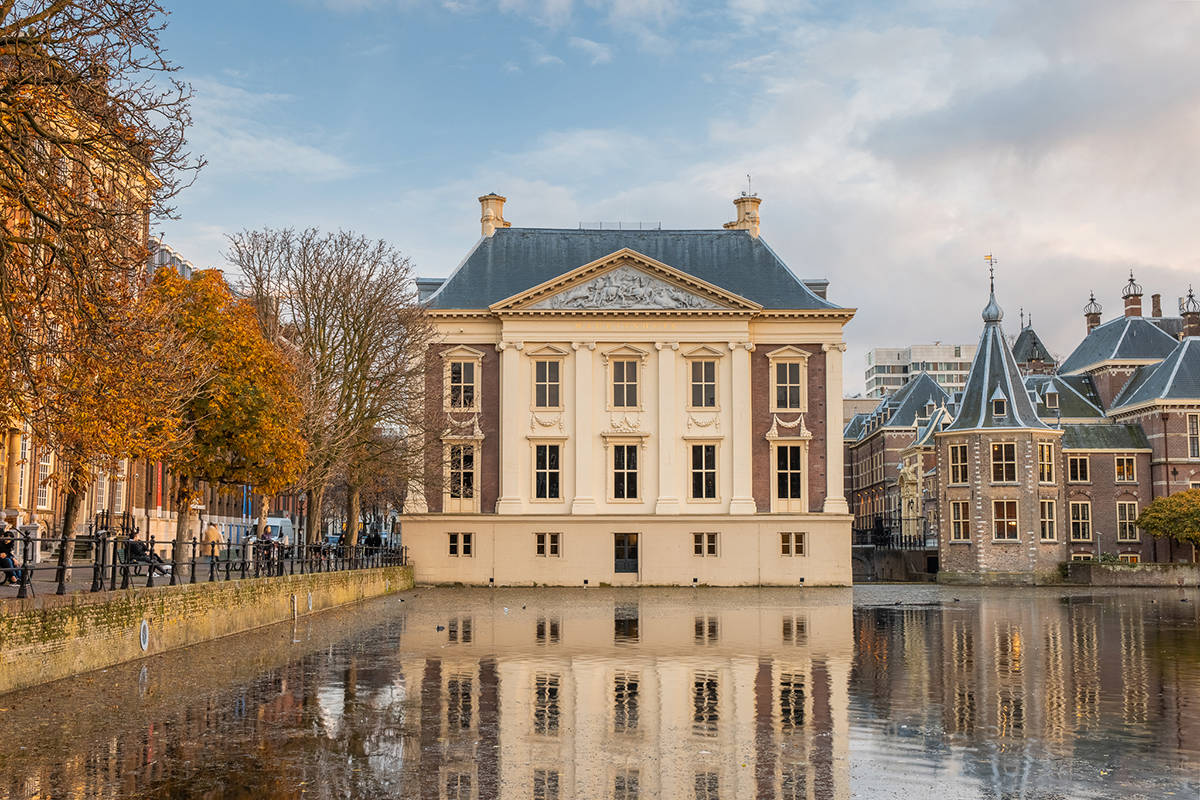 A cream colored building behind a small body of water in the foreground. The reflecting image of the building is visible on the water.