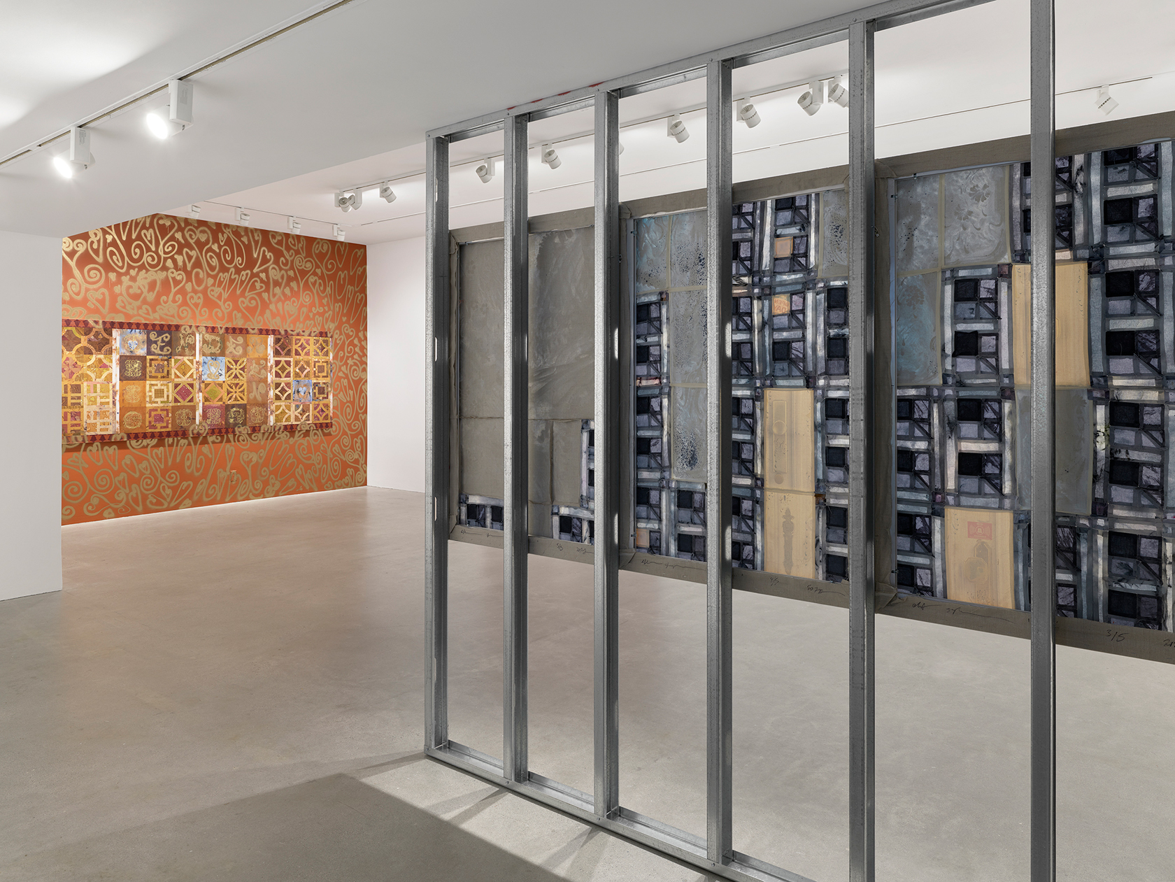 A gallery entryway with scaffolding on the right and the back of a blue patchwork-style painting in view. In the background, a painting hangs on an orange wall adorned with gold spray-painted motifs.