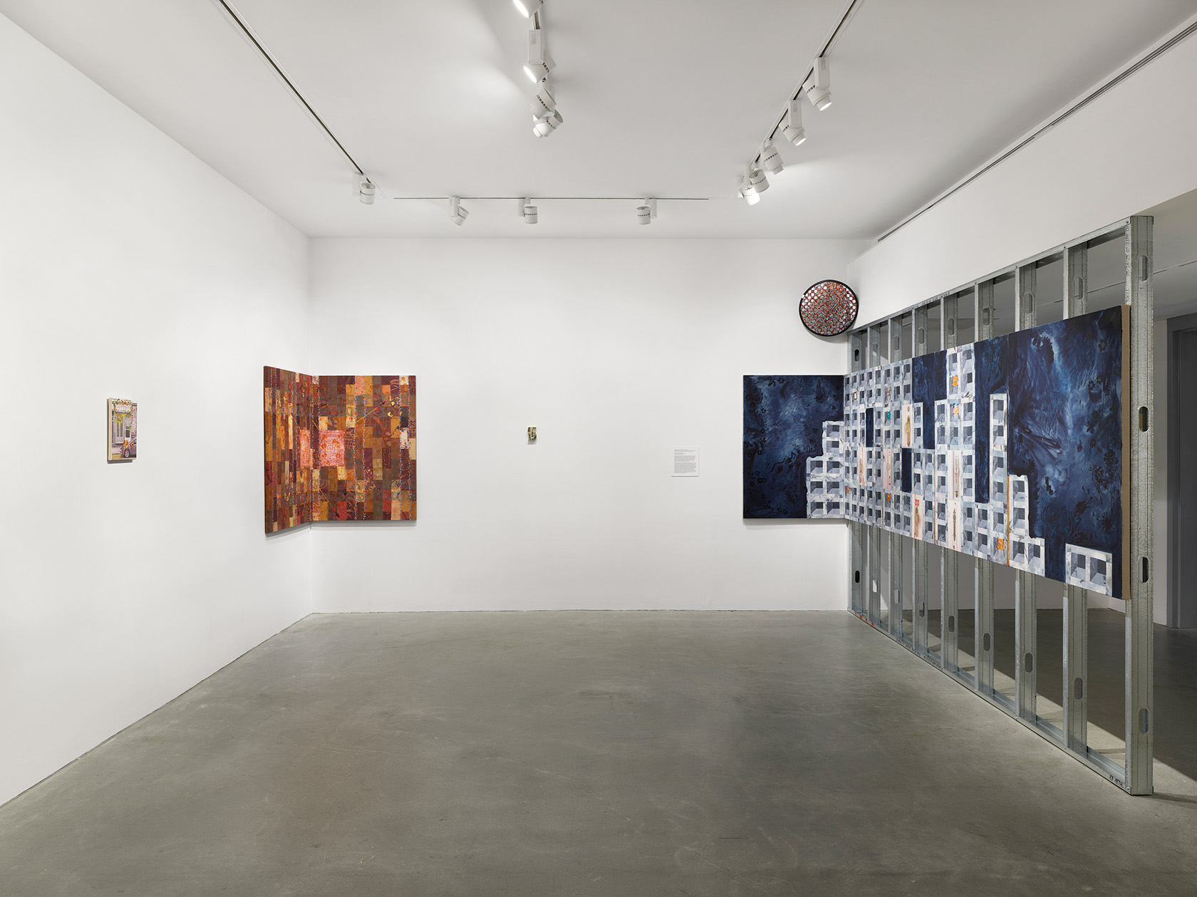 A blue cinder block-patterned painting wraps from scaffolding to the wall; a painted mirror and three other paintings hang throughout the gallery.