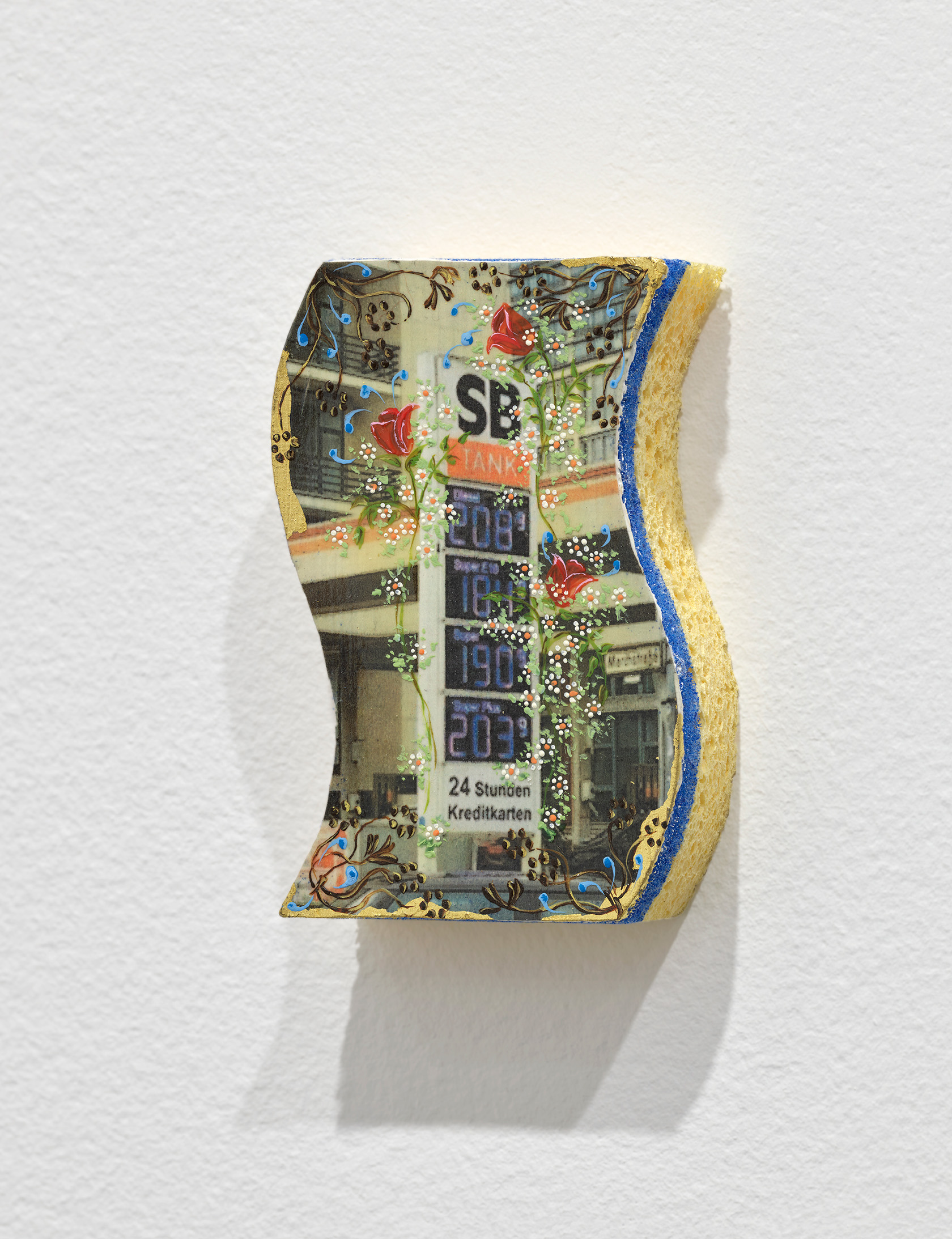 One side of a yellow and blue kitchen sponge is painted or printed with a view of a gas station. This scene is adorned with delicately painted flowers and other ornamental gestures.