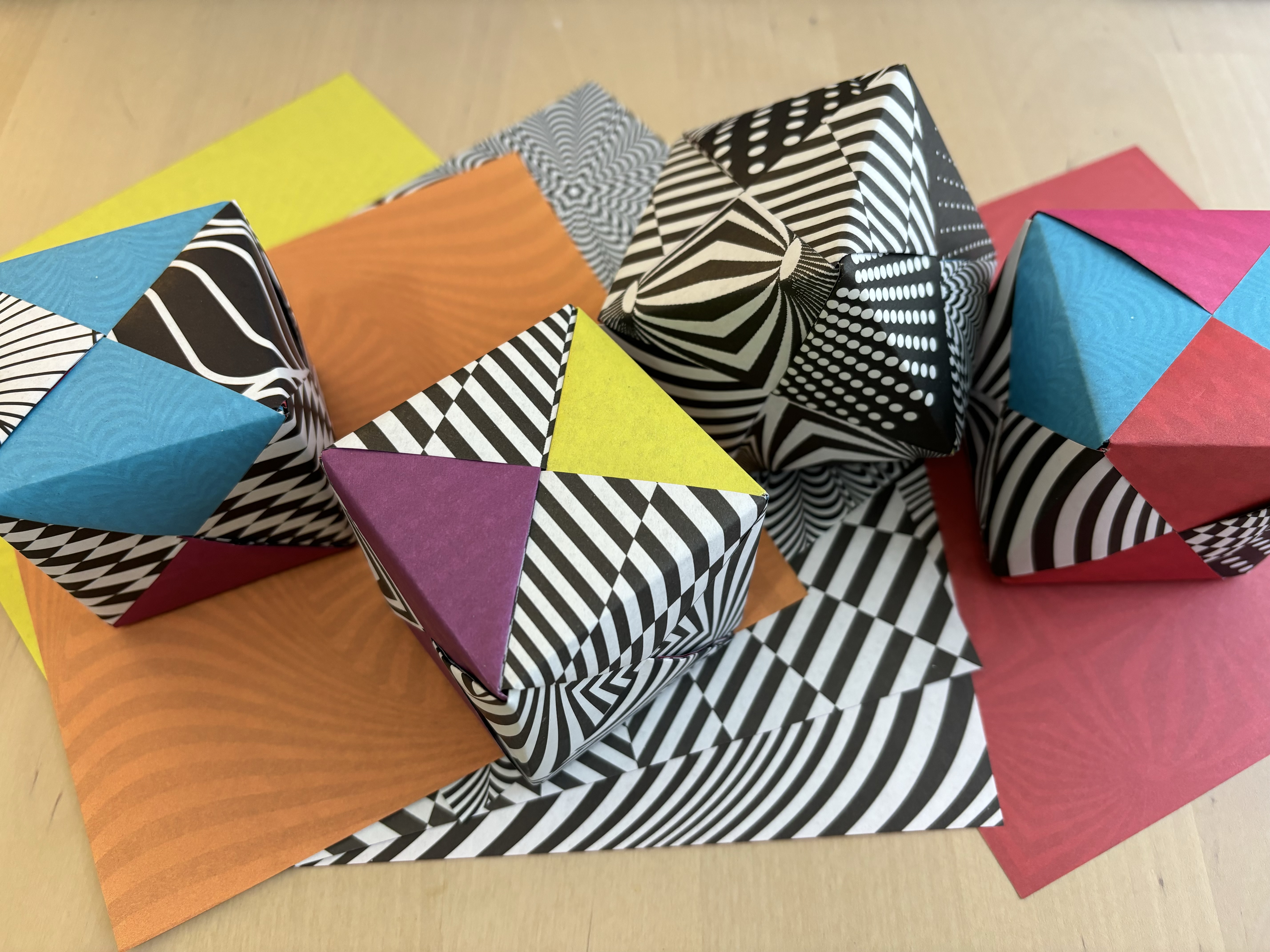 Aerial view of four cubes made out of origami paper sitting on a pile of origami paper on a light wood table.