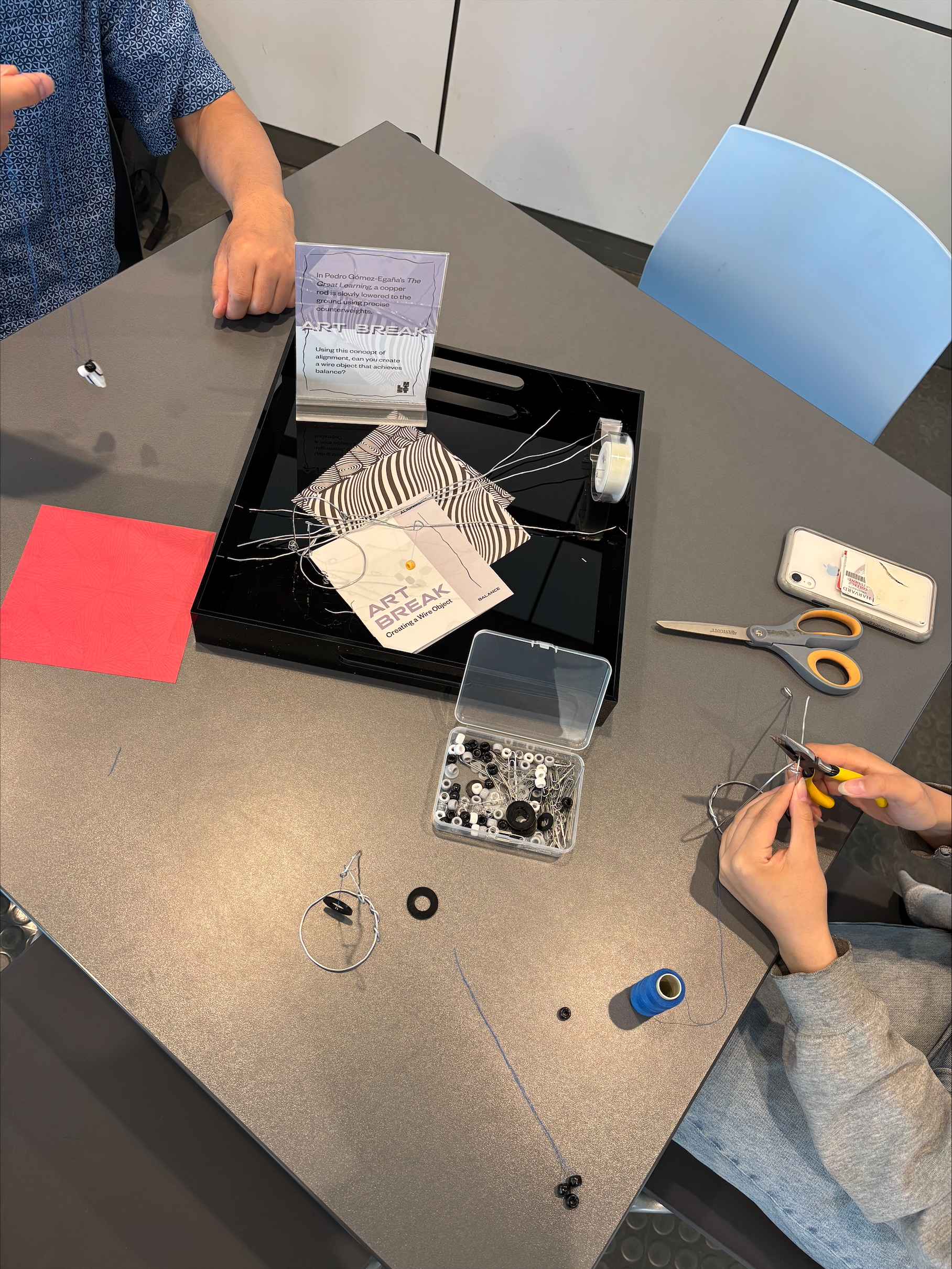 Aerial view showing the hands of two people making kinetic sculptures out of wire and beads. There is a black tray with more supplies on the table in between the two people.