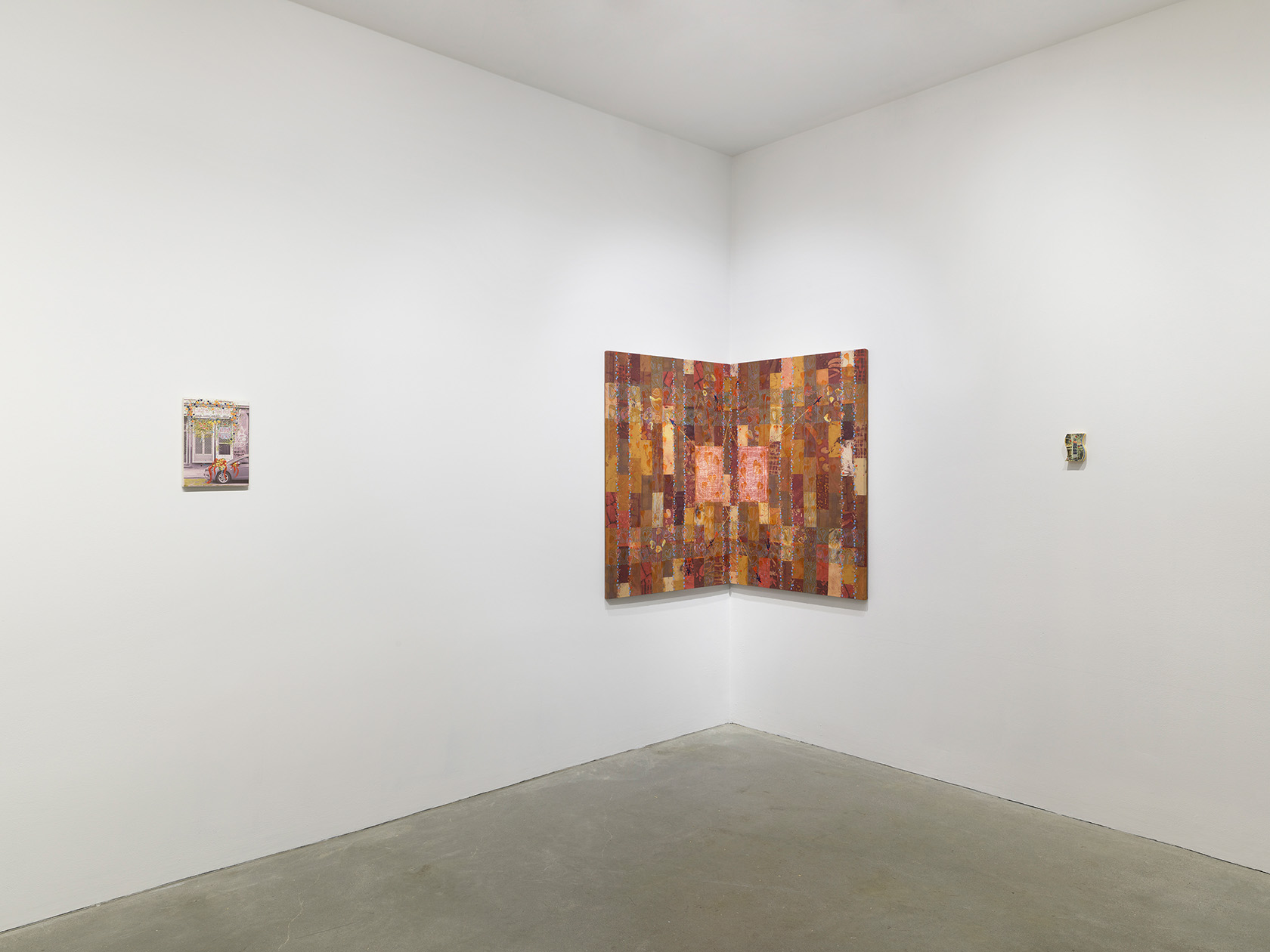 A corner of a white-walled gallery where three paintings are on display. On the left is a small painting of a street scene. In the middle, a painting featuring a brick-like pattern in shades of red, brown, pink, and orange is positioned in the corner of the room. On the right, a small painting on a sponge is mounted on the wall.