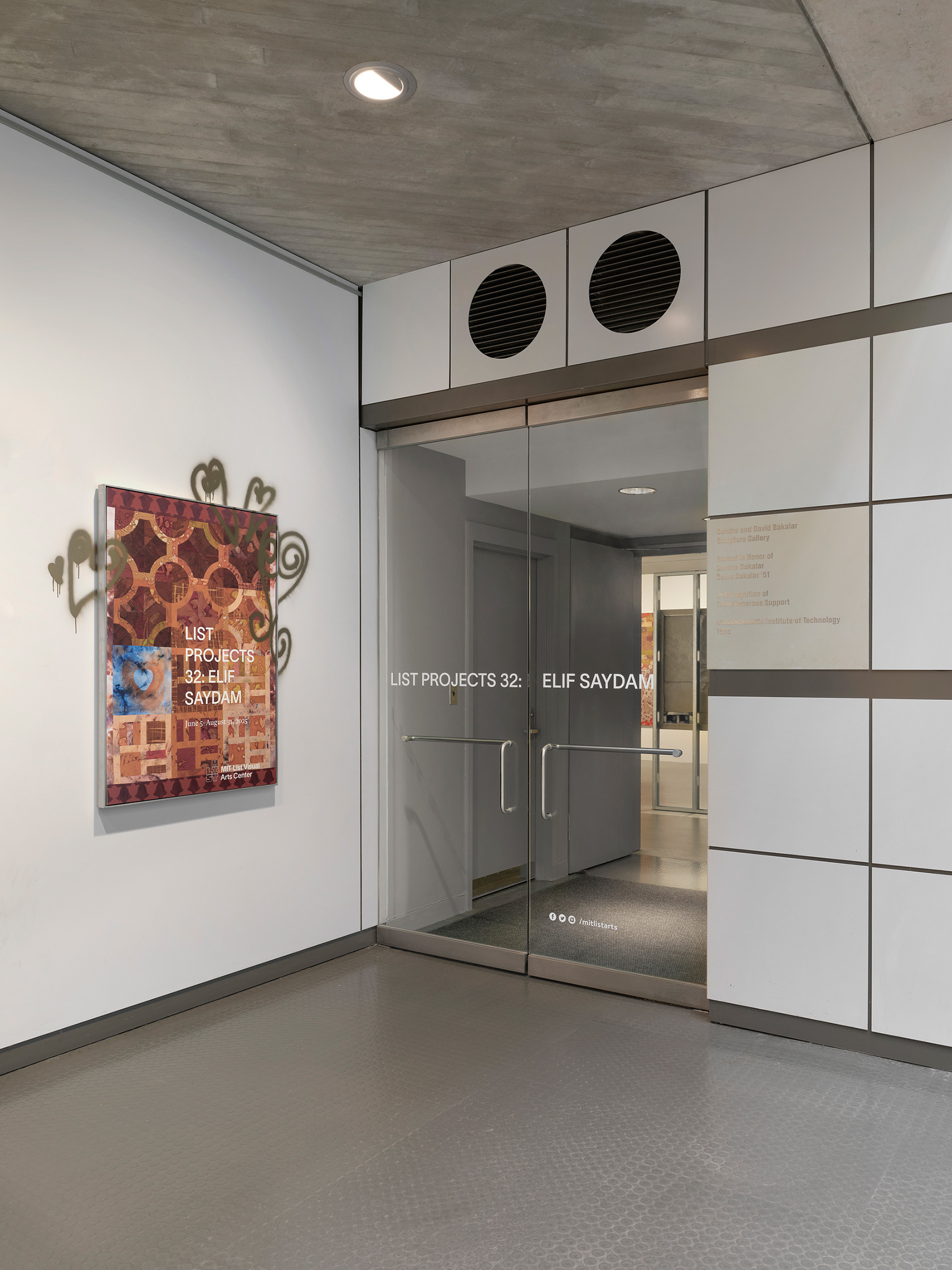 An entryway to a gallery with glass doors. Text reading “List Projects 32: Elif Saydam” is displayed across the two doors. To the left of the entryway, a poster bearing the exhibition title hangs. The poster features a detail of a tile-like painting with gold spray-painted heart motifs that extend onto the adjacent white wall.