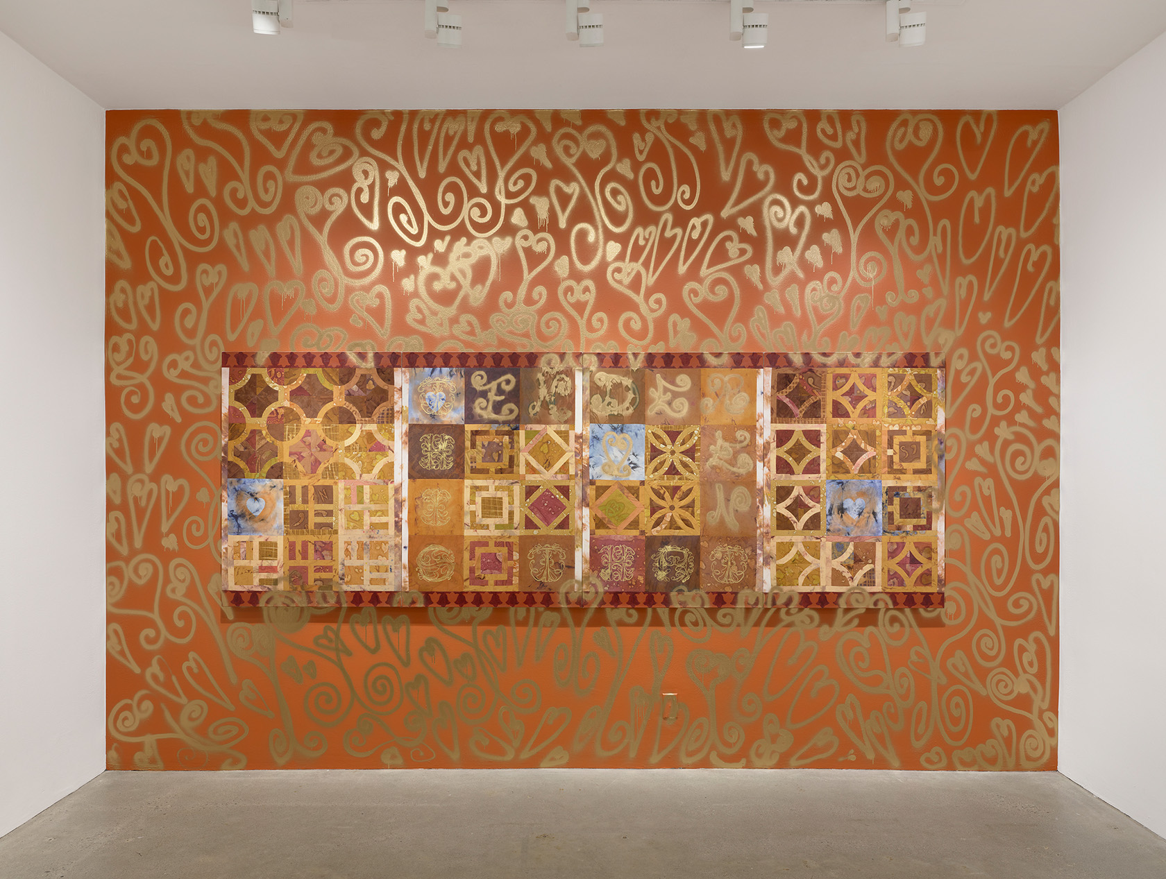 A painting with geometric breezeblock patterns in various shades of red and brown hangs on a rust-colored wall. Gold spray paint in exuberant heart shapes covers the surrounding wall, bleeding onto the wall and onto the canvas itself.